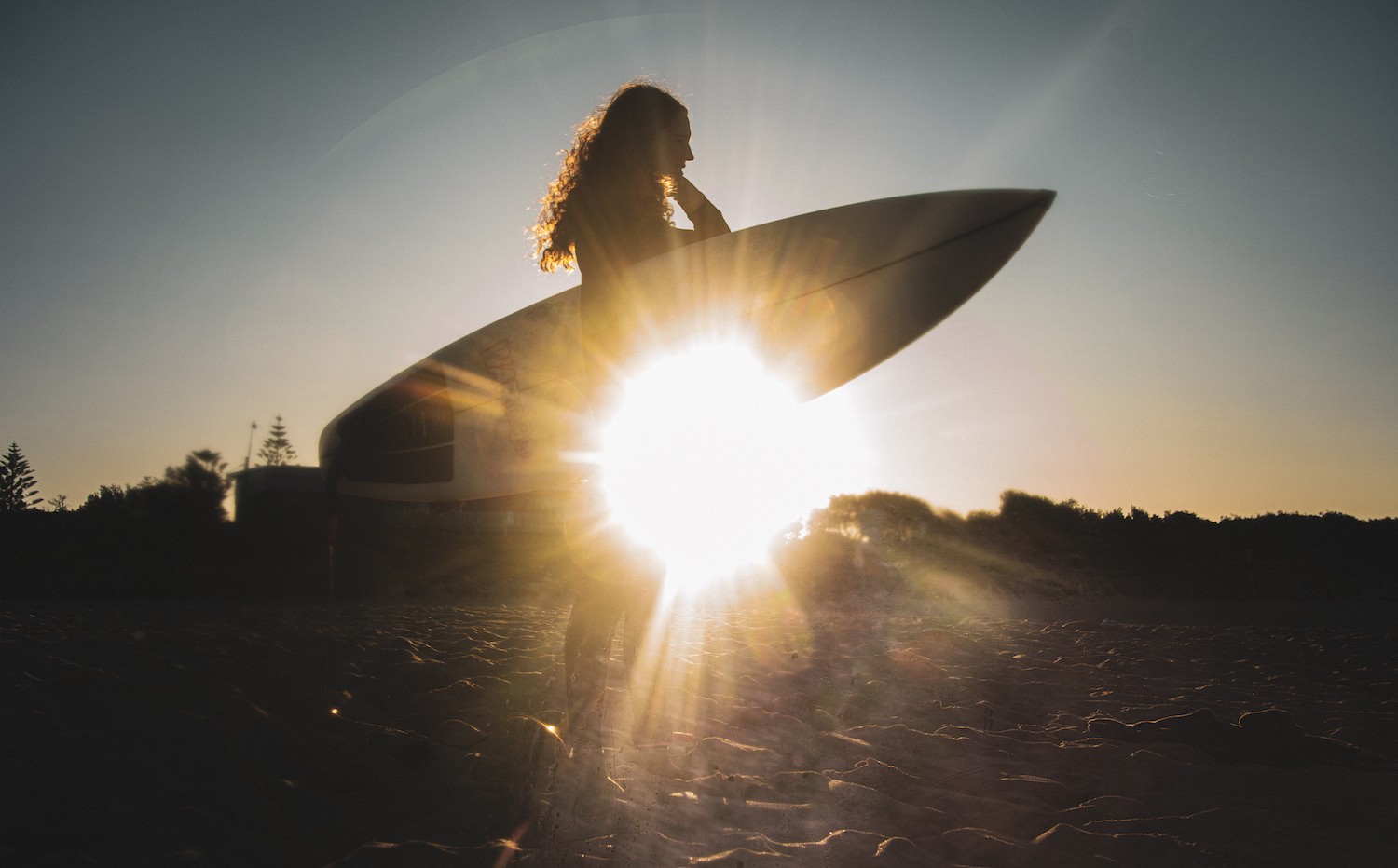 Silhouette of woman holding surfboard against sun on horizon