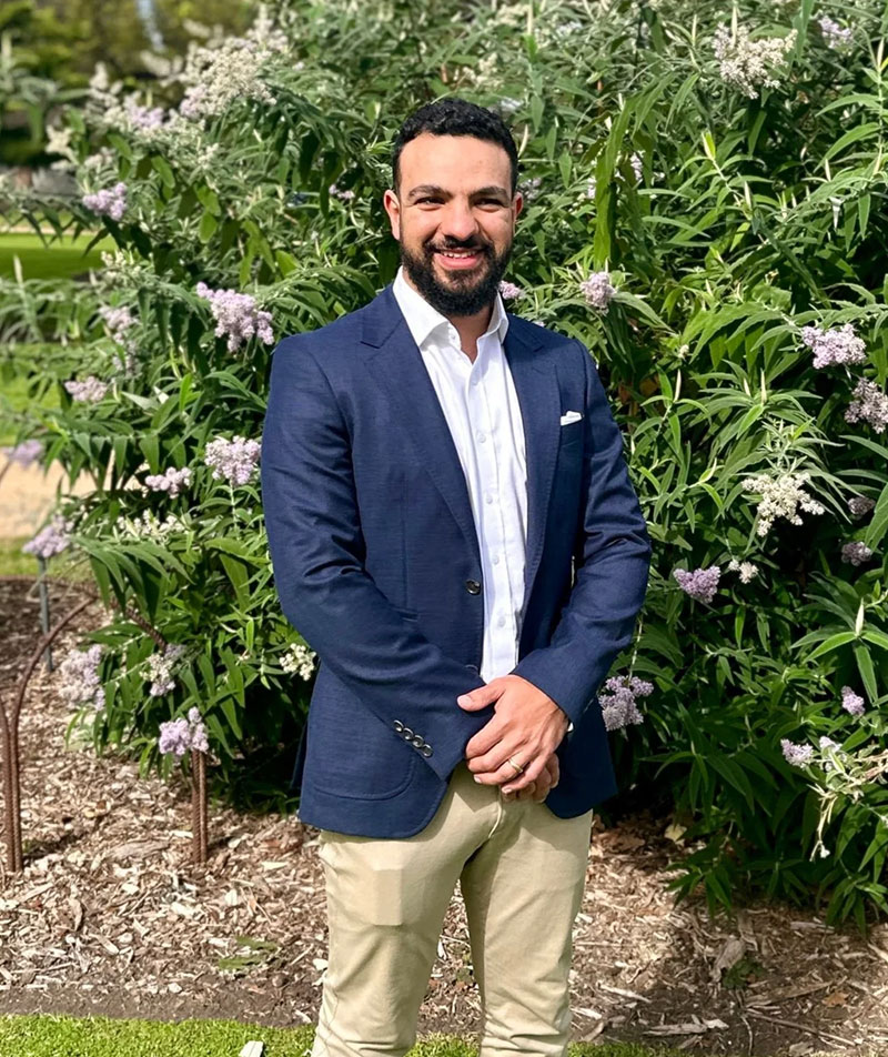 A profile image of Meena Iskandar in a professional outfit in a leafy outdoor setting