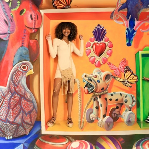 A woman stands inside a colorful, surrealist mural featuring various animal motifs and vibrant butterflies.