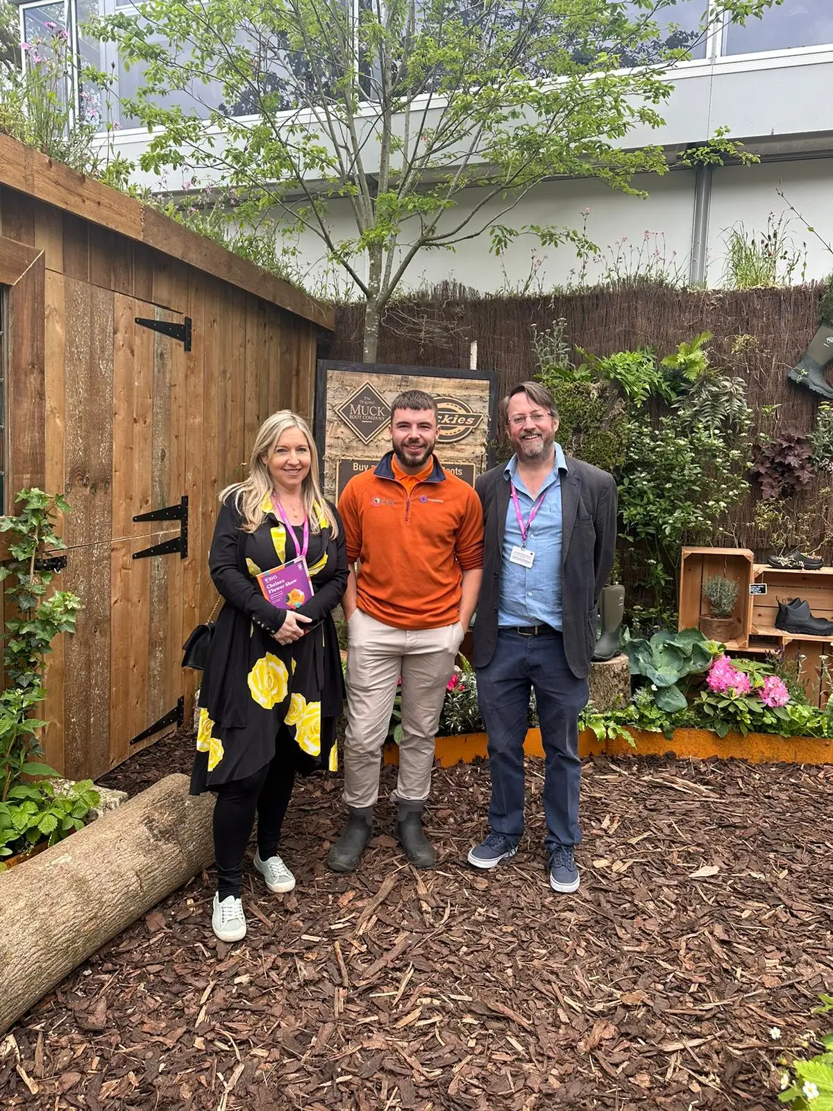 Three people stand together in a garden, surrounded by plants and wooden structures, smiling for the photo.