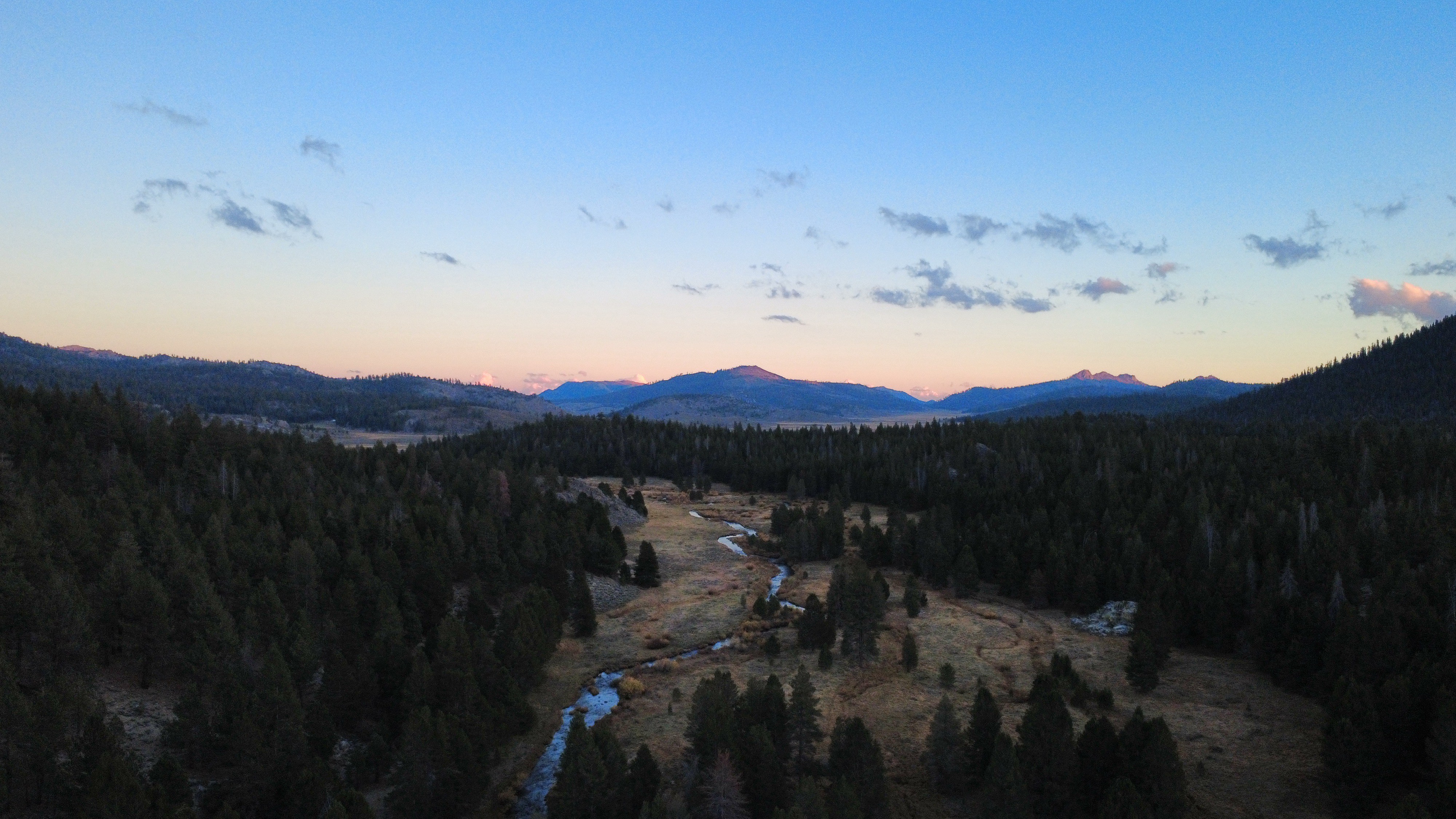 A winding river flows through a forested valley at dusk with mountains in the background under a clear sky.