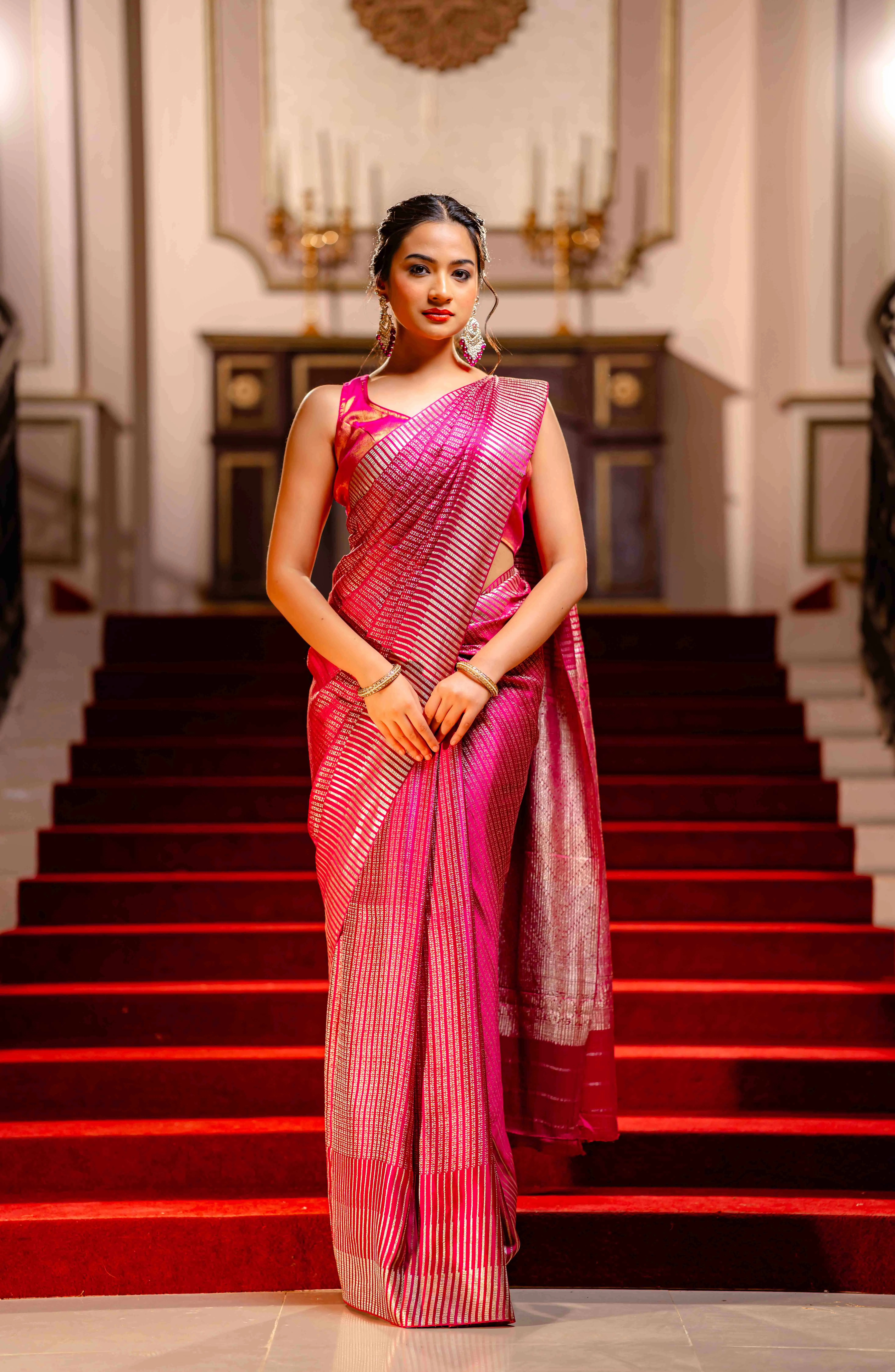 A woman wearing a red saree with golden borders from That Saree Shop, seated gracefully on a chair in an elegant room, highlighting the saree’s luxurious weave and classic festive charm.