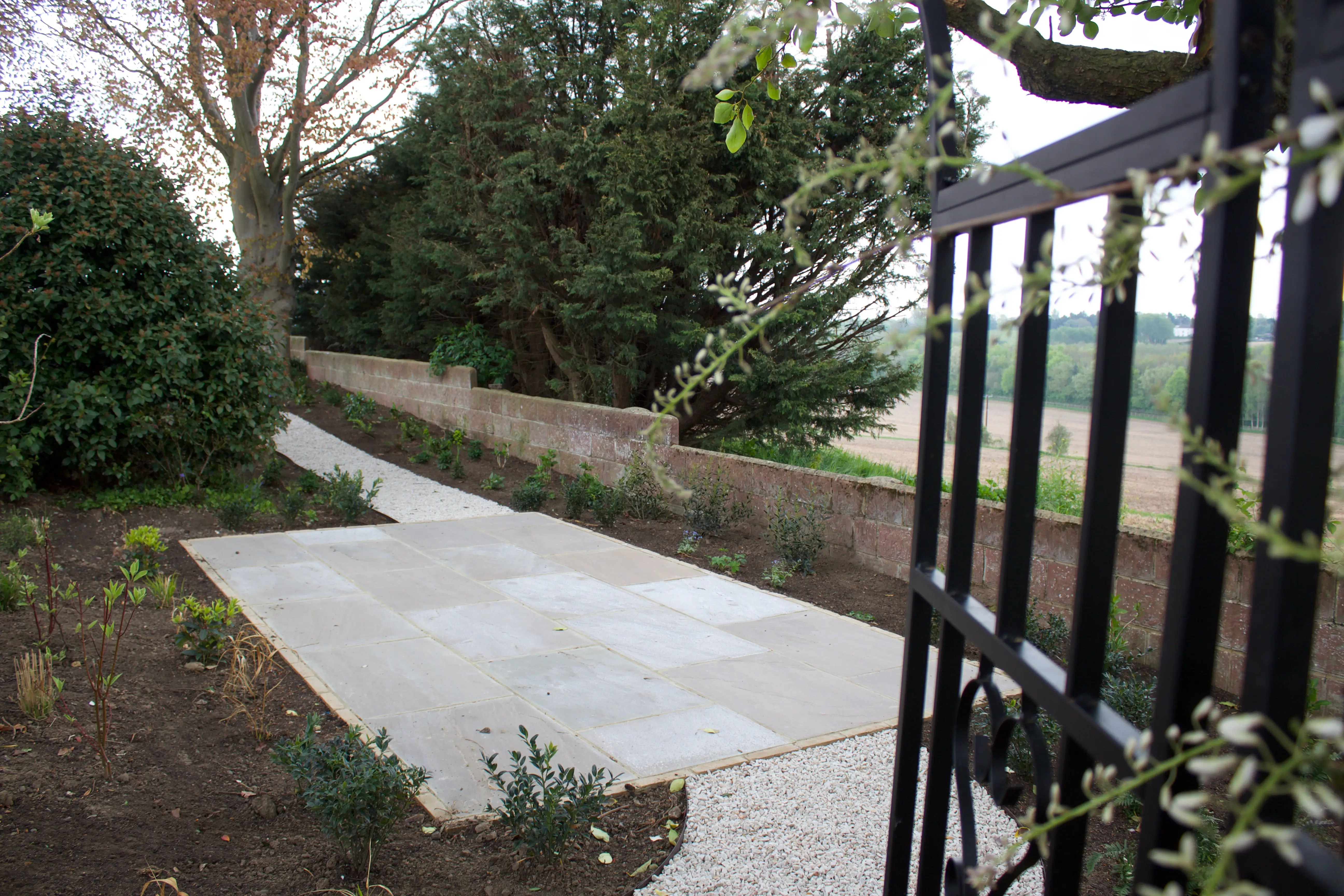 A pathway lined with greenery, viewed through a black metal gate, leading to a quiet outdoor area.