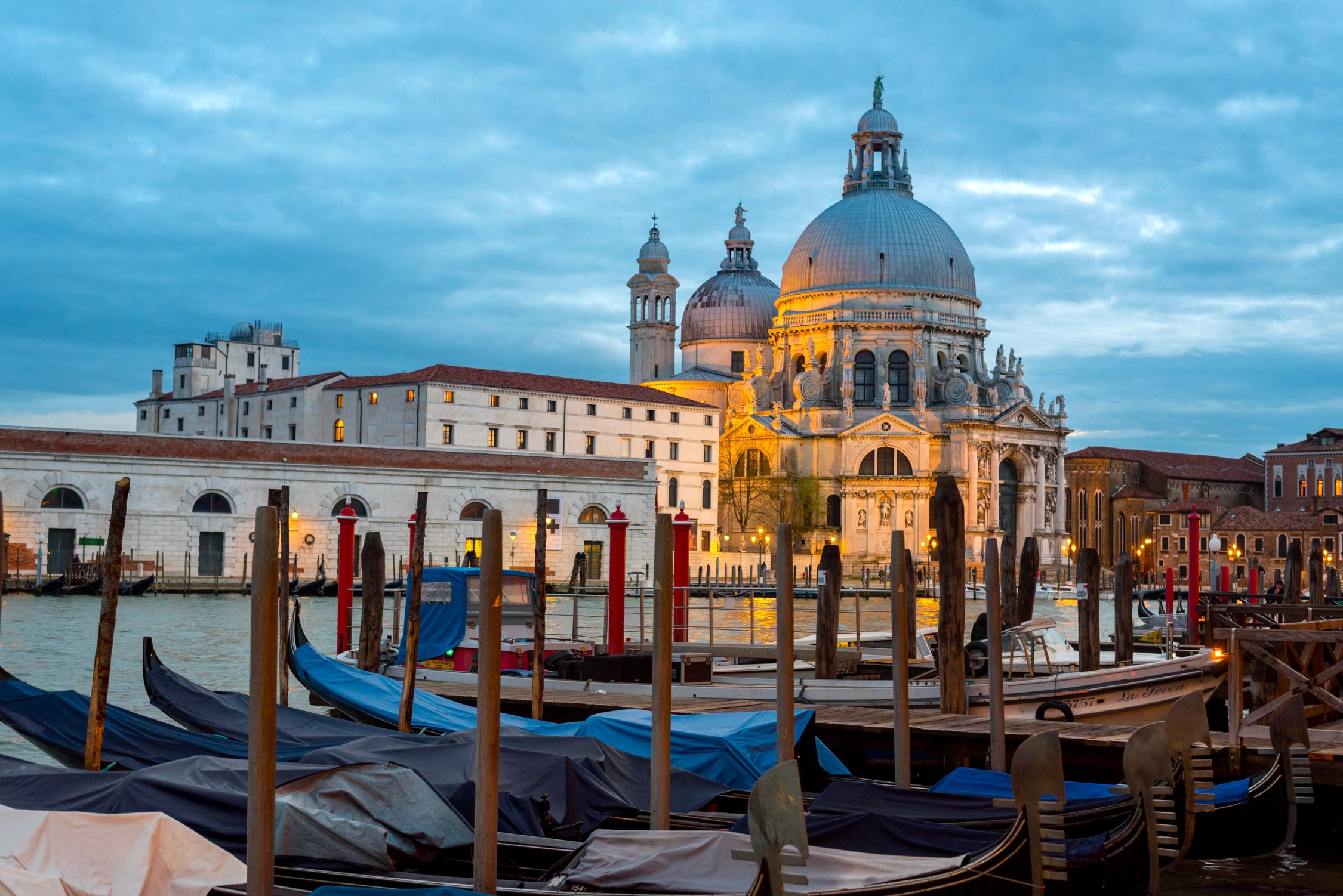 Rows of colorful gondolas moored in a canal in front of a large domed church under a blue sky at dusk.