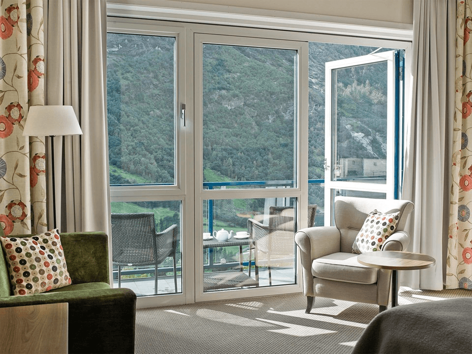 A cozy hotel room with a chair, small table, and floral patterned curtains opens to a balcony overlooking lush green mountains.