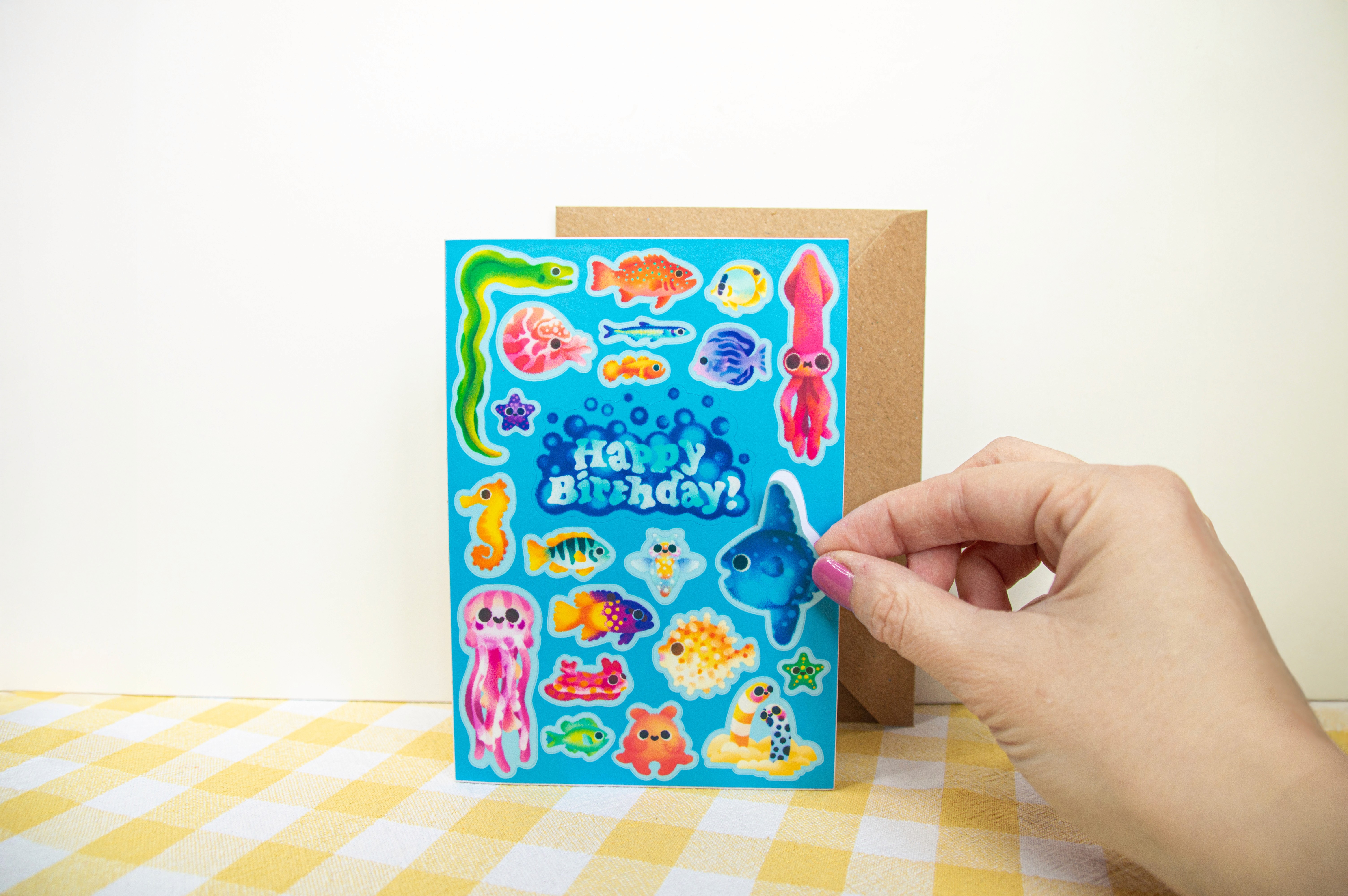 A sky blue card that is also a sticker sheet. It is ocean creature themed and a hand is peeling off a sticker of a sunfish. The centre of the card has a bubbling "Happy Birthday" sticker.