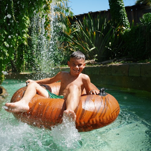 A boy enjoys a ride on an inflatable tube in a water park, surrounded by greenery and a cascading waterfall.