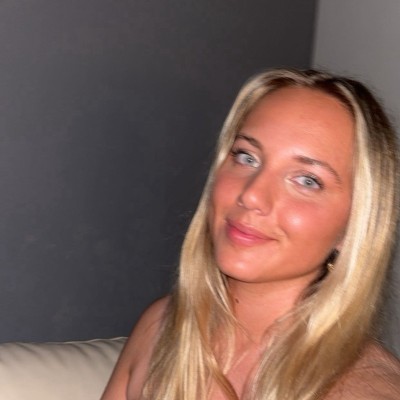 A woman with long hair, wearing sunglasses and smiling on a sunny street with trees in the distance.