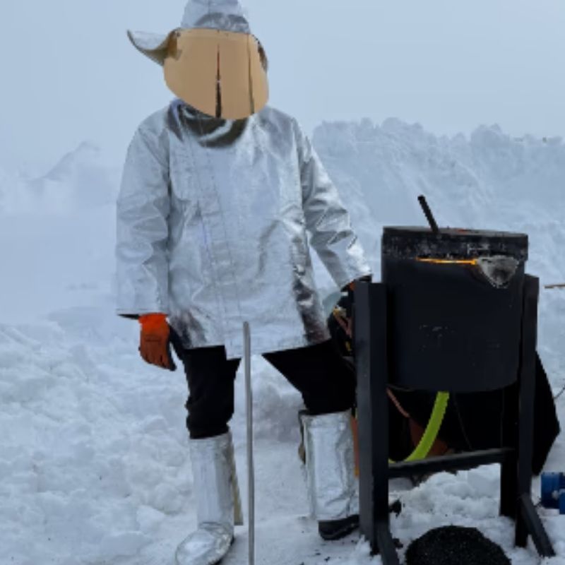 Zwei Mitglieder des Lava Show-Teams, gekleidet in reflektierende hitzeschützende Anzüge und Helme, sind zu sehen, wie sie Lava auf dem Langjökull-Gletscher in Island während einer privaten Veranstaltung schmelzen. Umgeben von Schnee und vereistem Gelände, bedienen sie die Lava-schmelzenden Geräte, wobei die glühende Lava sichtbar in den Behältern leuchtet. Der auffällige Kontrast zwischen der feurigen Lava und der schneebedeckten Landschaft hebt die dramatische Verbindung von Feuer und Eis hervor.
