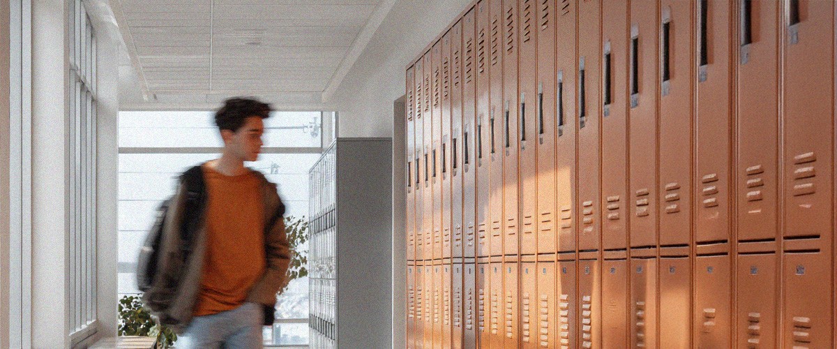 A young person with short, curly hair sits at a desk working on a computer in a bright classroom or office setting. They appear focused on the screen, with other people and desks softly blurred in the background, suggesting a collaborative learning or work environment.