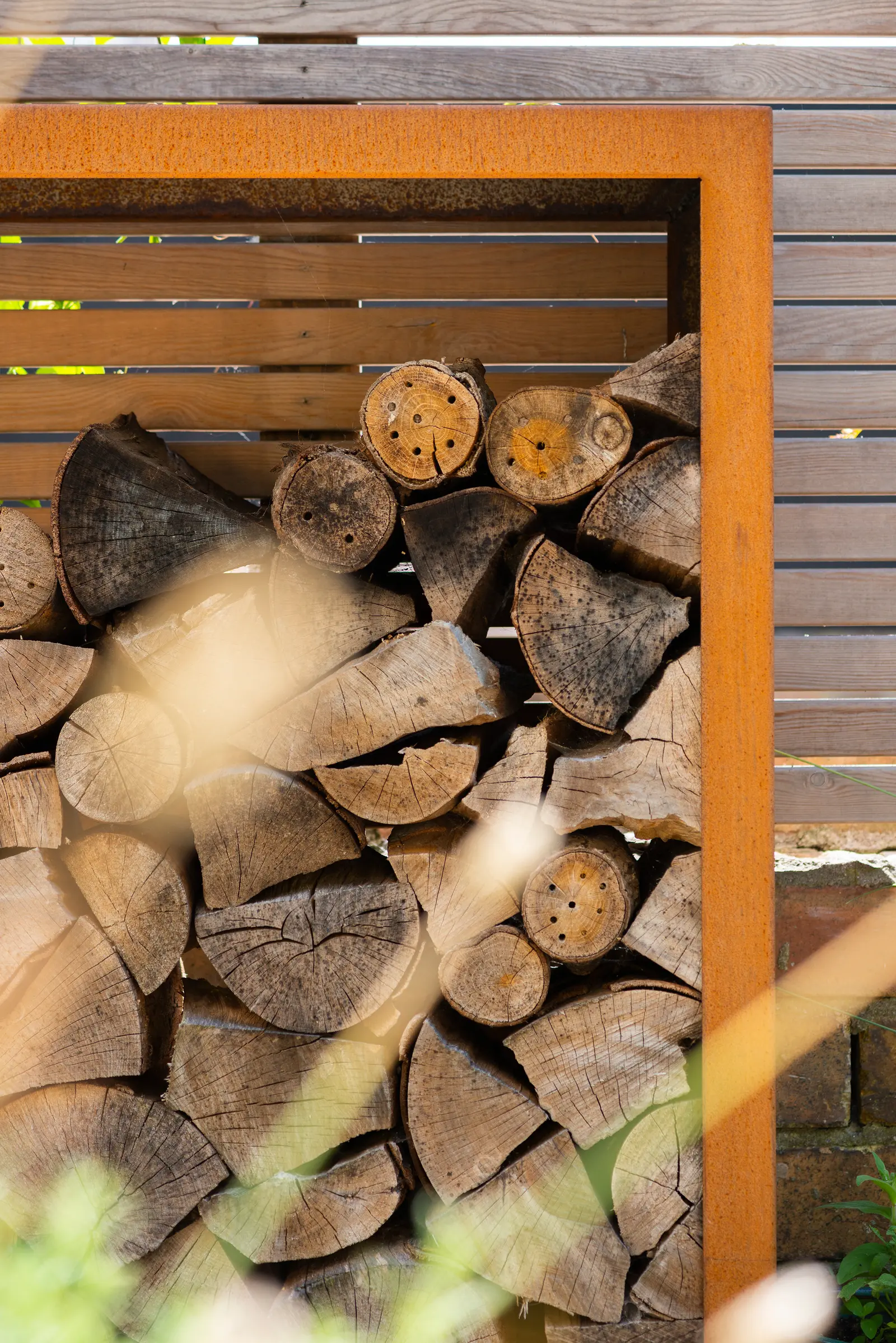 A stack of neatly arranged firewood is visible in a wooden shed, surrounded by green grass and plants.