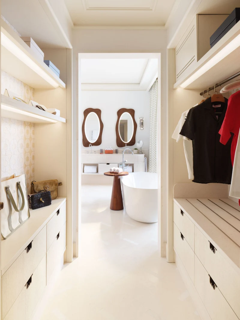 Ein Blick durch einen begehbaren Kleiderschrank mit hellen Einbauschränken, beleuchteten Regalen und Handtaschen auf ein Badezimmer mit einer freistehenden Badewanne und zwei Spiegeln.