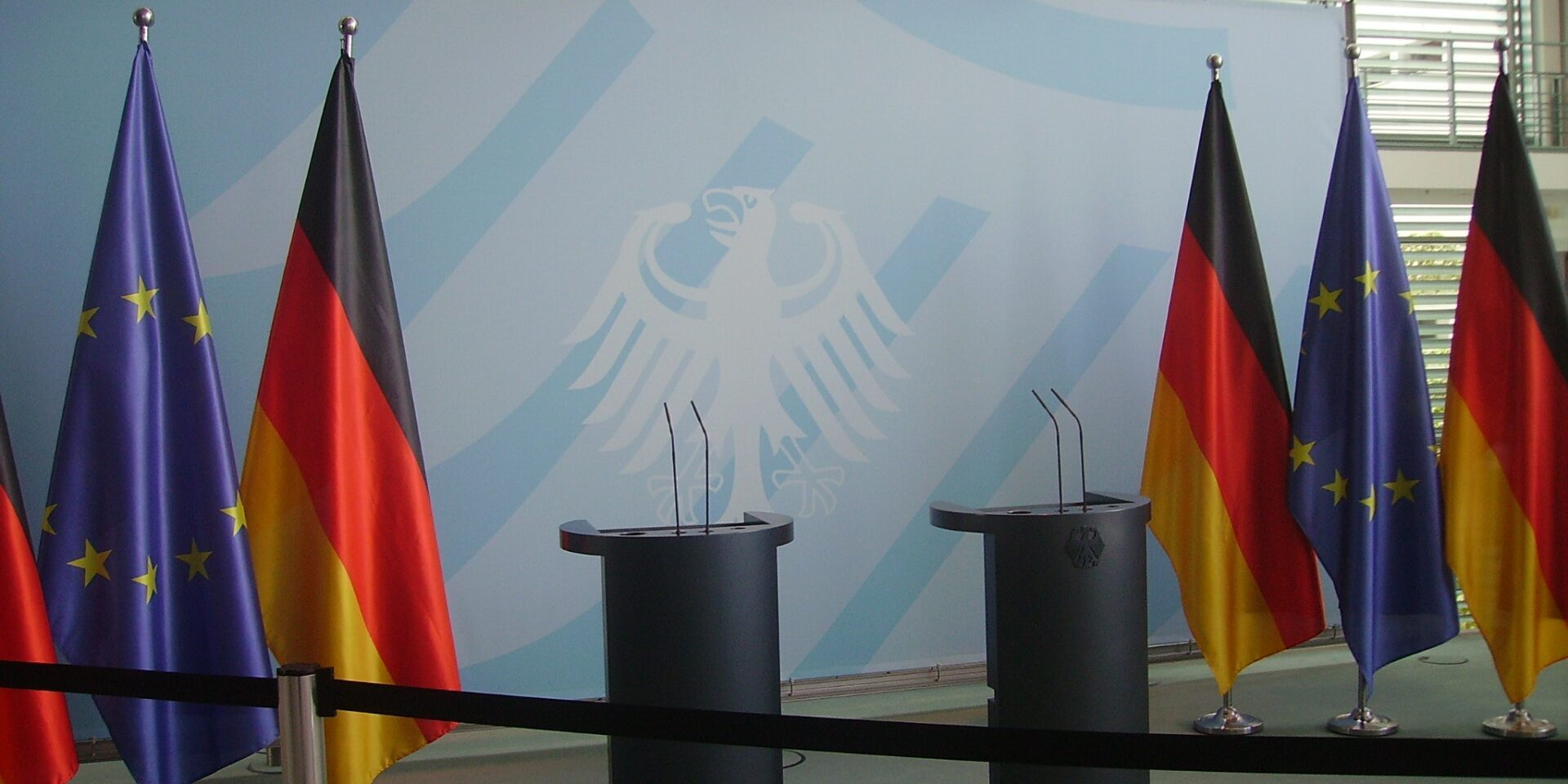 The image shows two podiums set up for a press conference with German and European Union flags in the background, highlighting an official diplomatic event.