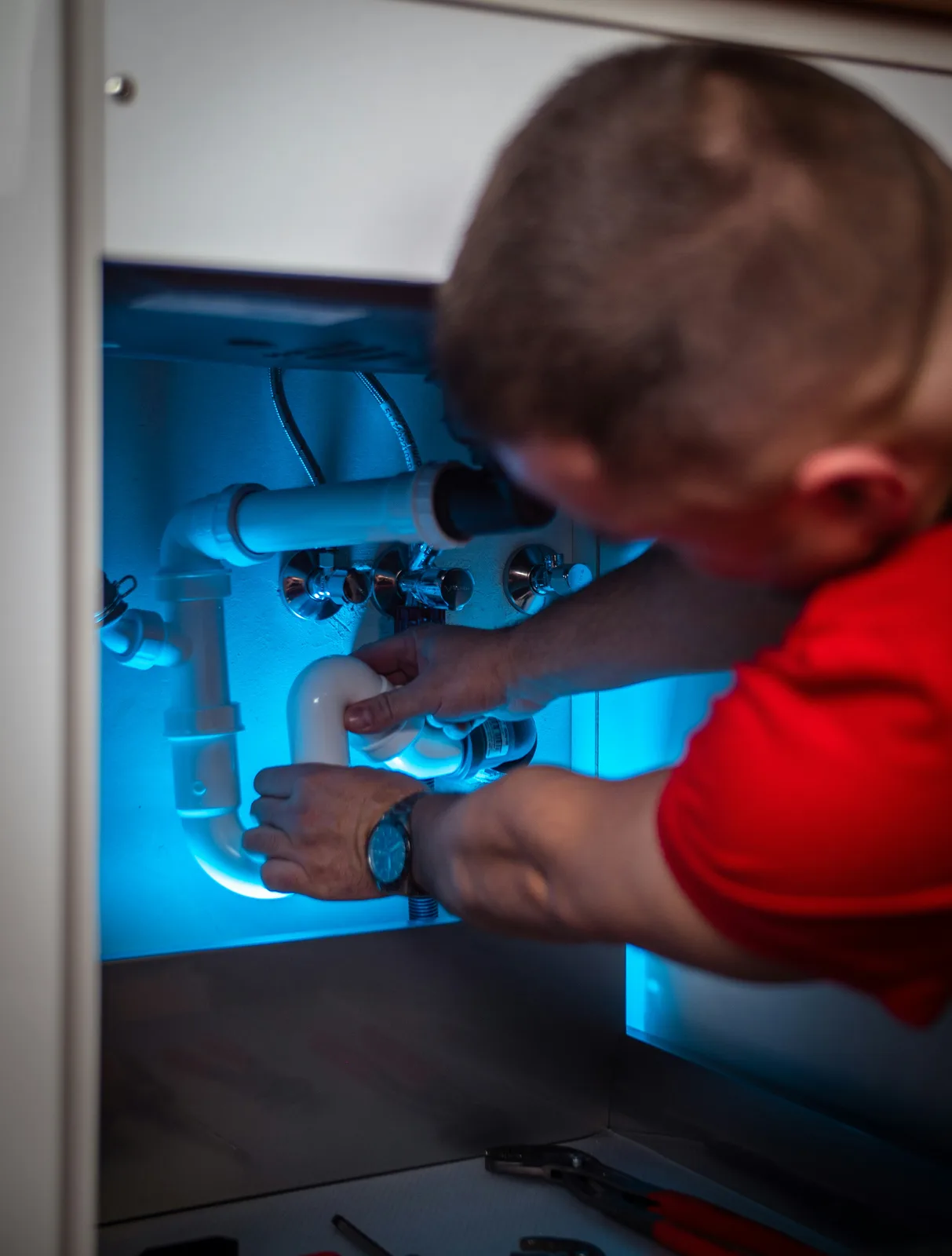 Man repairing sink drain pipes under cabinet with blue backlighting
