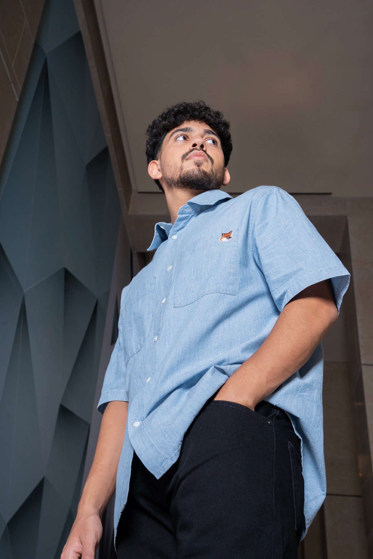 A young man posing against an angular teal wall in a light blue short-sleeve shirt and black trousers.