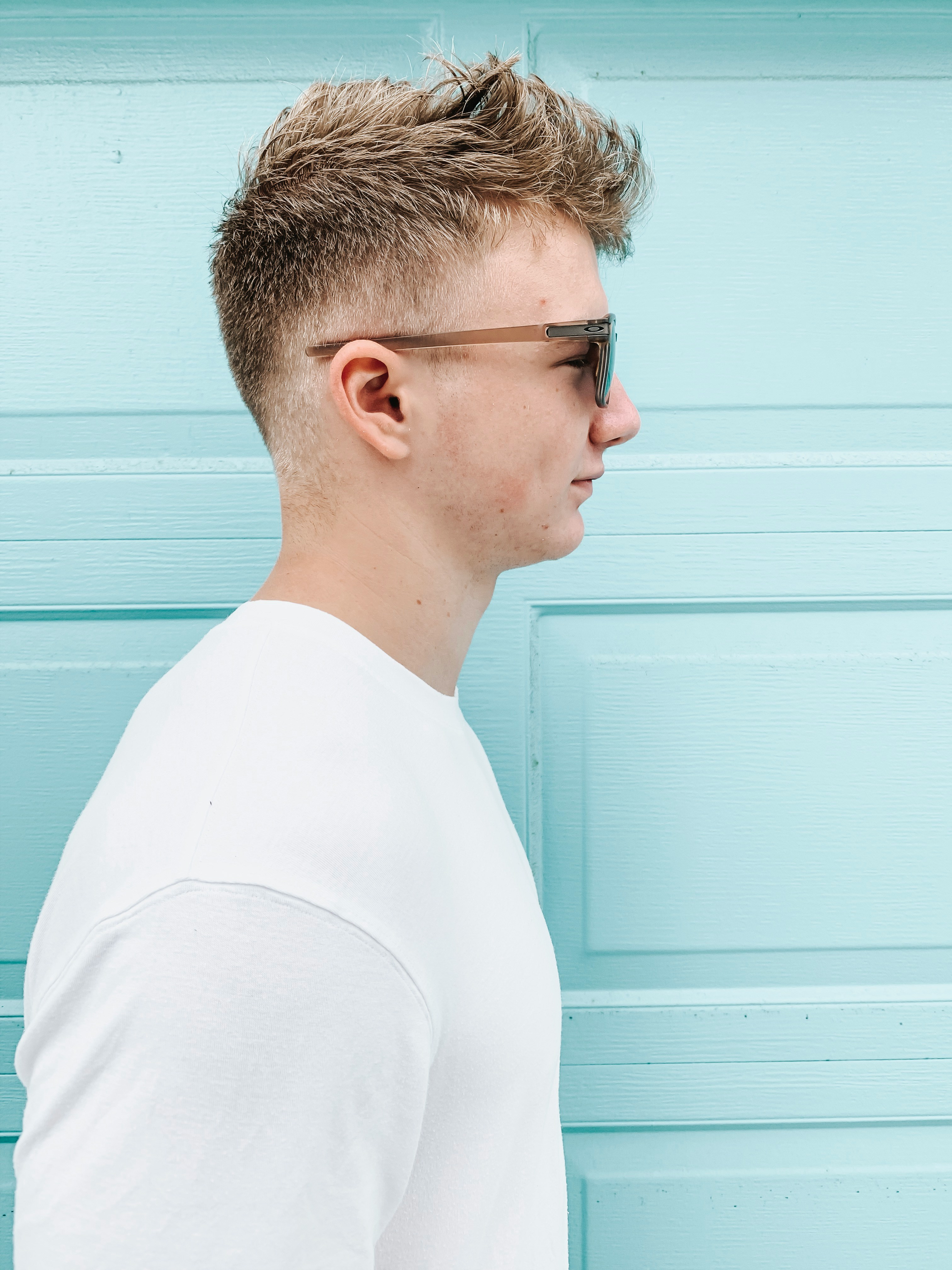 A young man with light brown hair wearing sunglasses and a white shirt against a blue background.