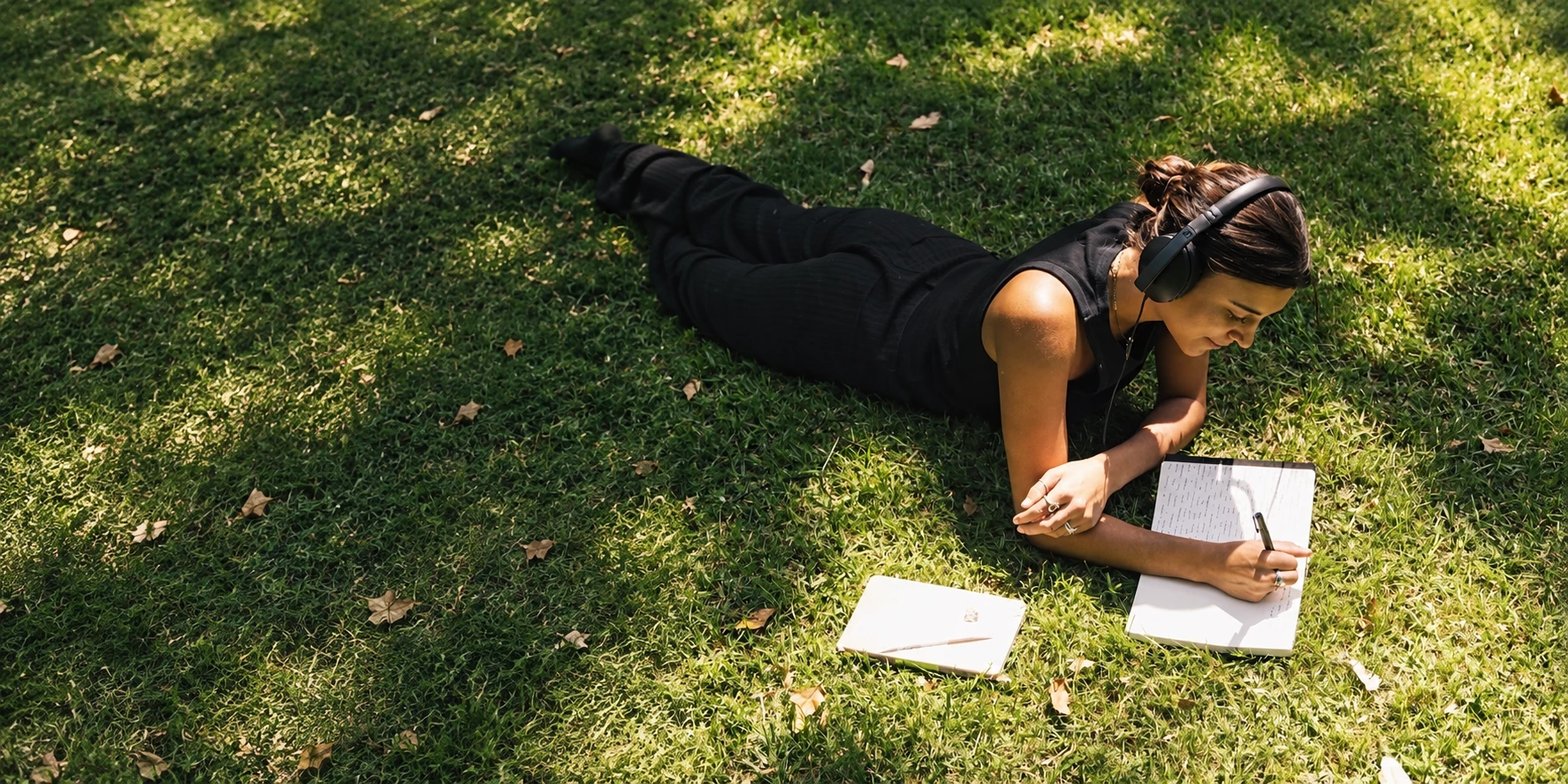 A woman wearing headphones and writing in a notebook on sunlit grass
