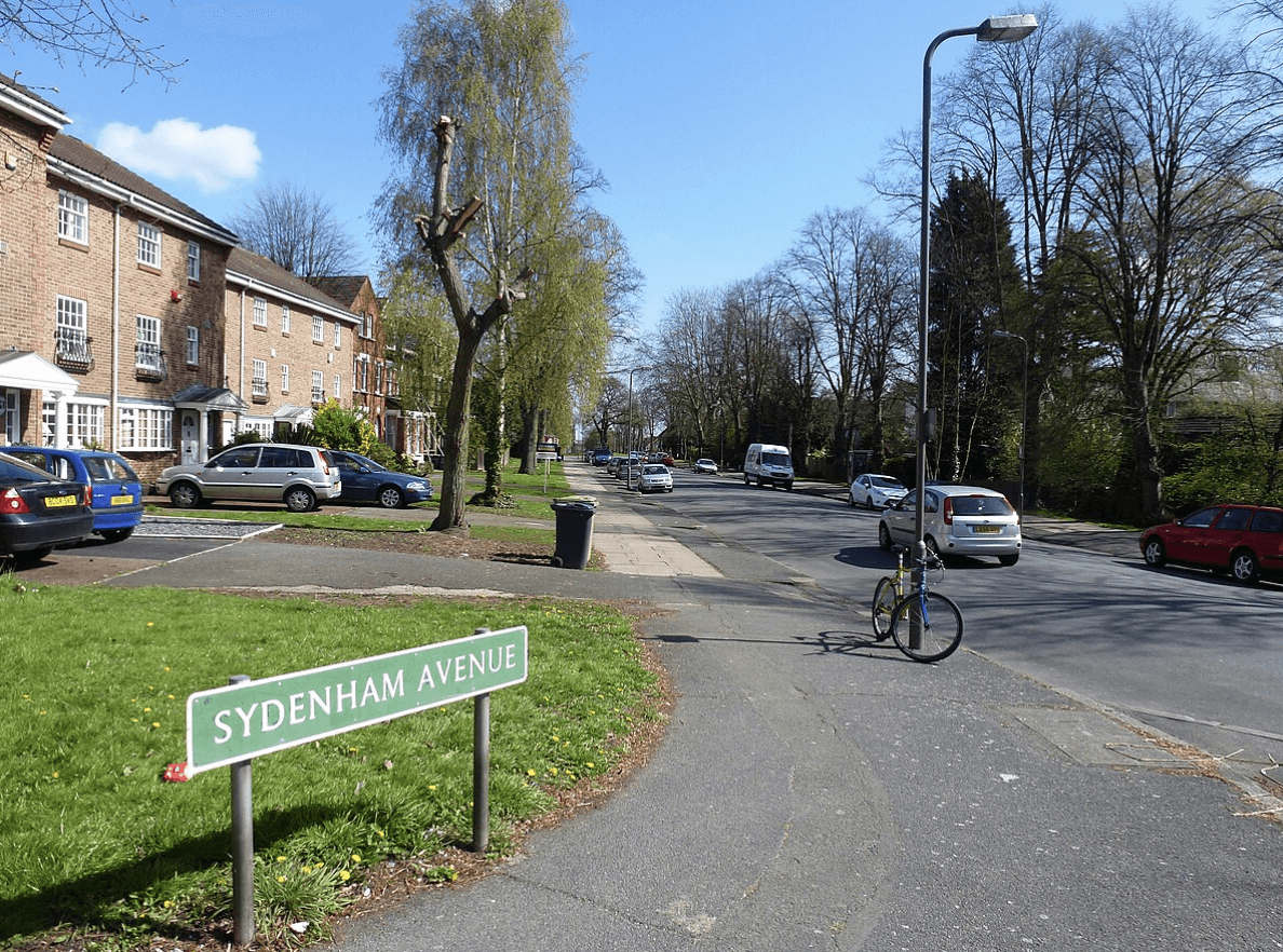 A smiling man with curly hair is sitting at a desk, looking at the camera, with a laptop and a lamp in the background.