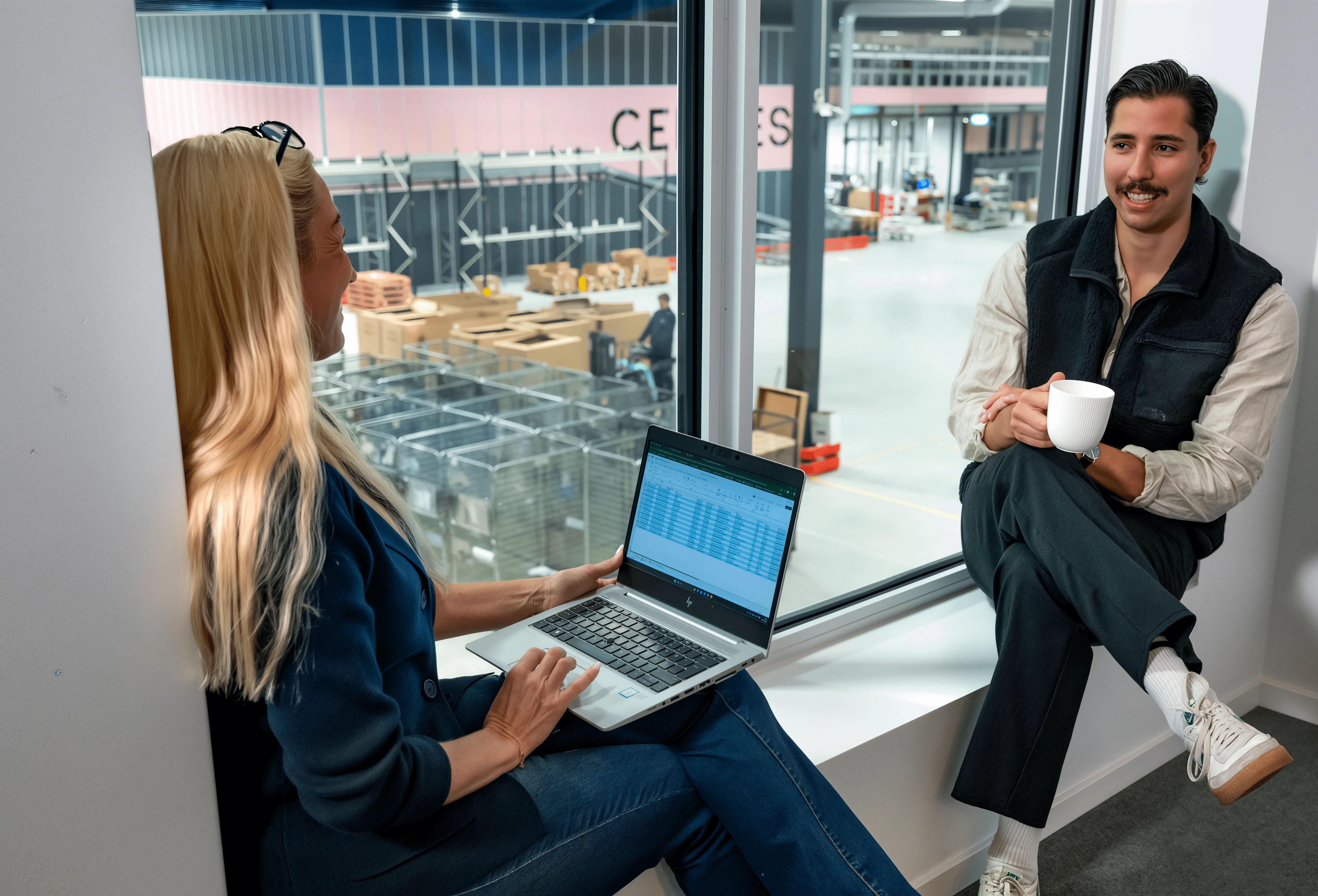 Kvinna och man diskuterar vid ett fönster. Kvinnan använder en bärbar dator. Mannen håller i en vit kaffekopp.