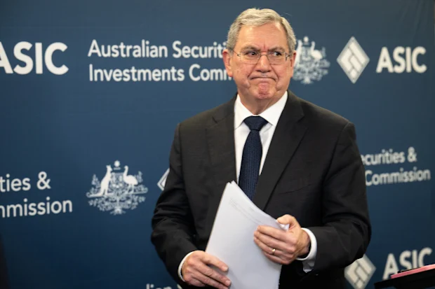 A man in a dark suit and glasses stands in front of an ASIC media backdrop holding papers, appearing serious during a press briefing