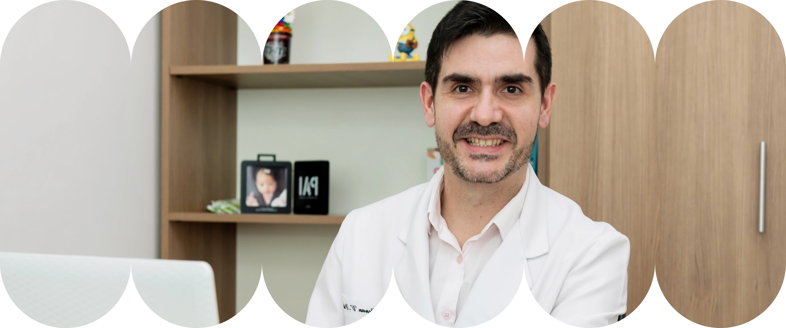 O Dr. Renan, médico de barba rala, usando jaleco branco sobre camisa rosa, sentado em uma mesa de escritório e digitando em um laptop. Prateleiras com decorações infantis, como Minions e um porta-retratos de "Pai", estão ao fundo.