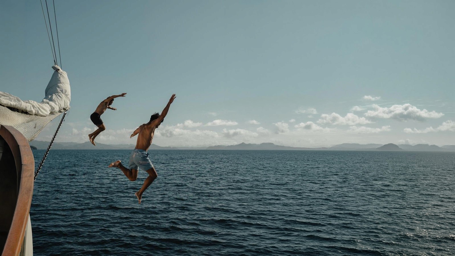 Two men jumping off a sailboat into open ocean water under a cloudy sky
