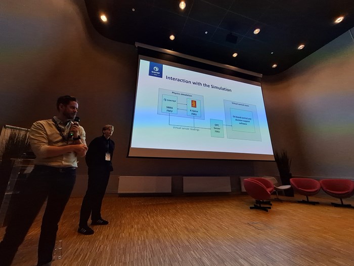 A presentation in a conference room with a large screen displaying graphs, and two speakers in the foreground.