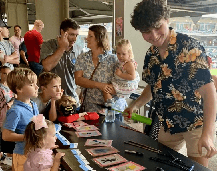 Group of children and parents gathered around a balloon artist creating designs at a table during an indoor event