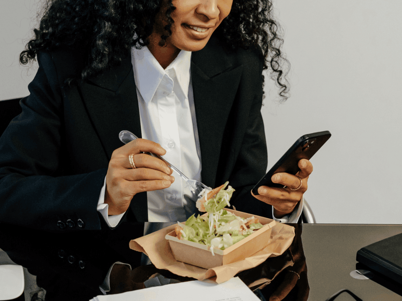 Businesswoman in a black suit enjoying a fresh salad while checking her smartphone, seated at a glossy black desk in a modern office setting.