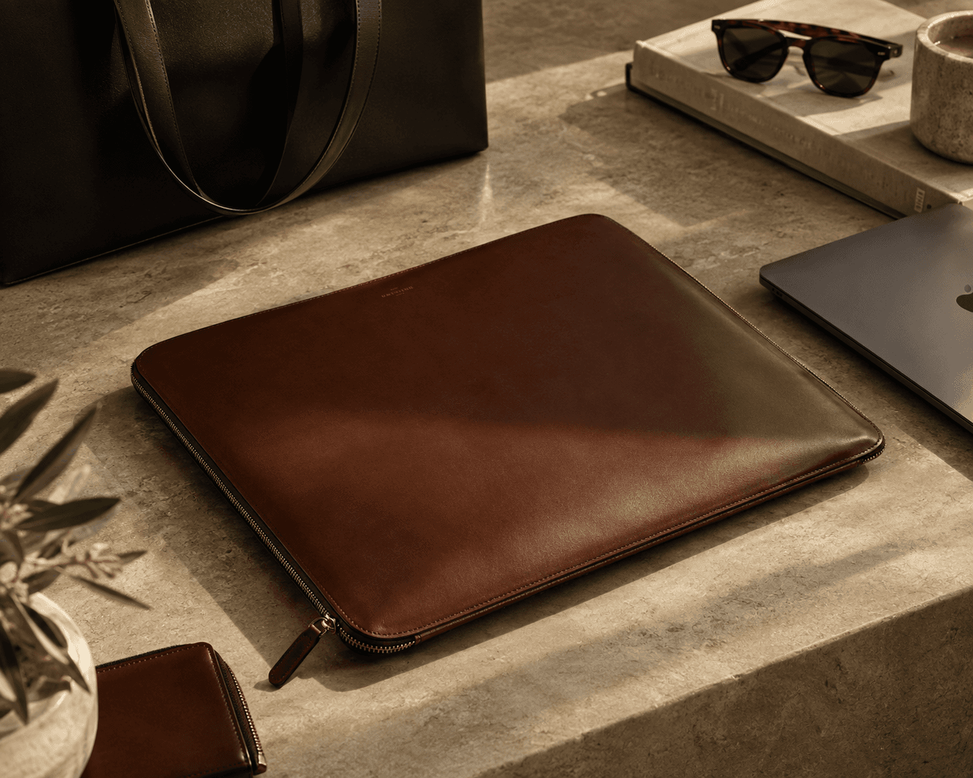 A refined tech and luxury accessory flatlay set on a sunlit beige travertine stone surface. At the center lies a smooth brown leather zip-around laptop sleeve. Surrounding items include a dark brown leather tote bag, a matching small leather wallet, a closed dark gray laptop, tortoiseshell sunglasses resting on a coffee table book, a ceramic espresso cup, and a potted olive plant casting soft shadows.