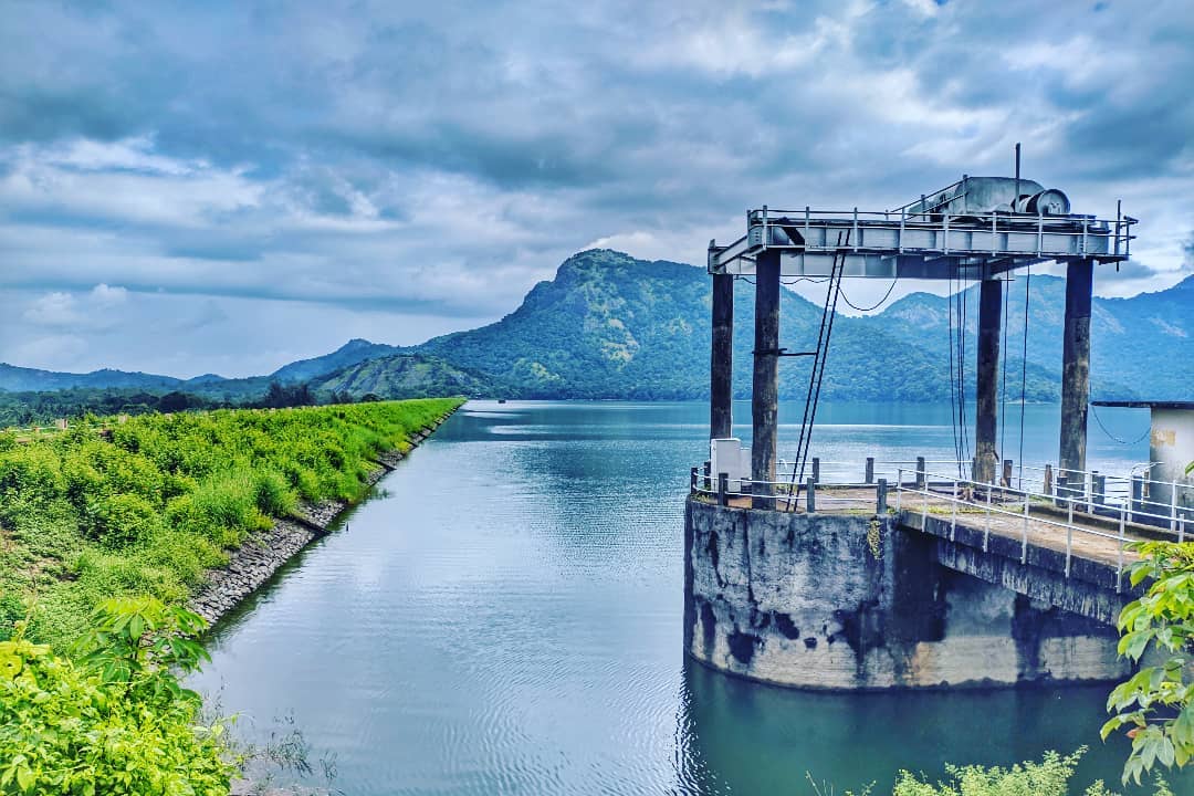 Scenic view of Pothundi Dam in Kerala, surrounded by lush greenery and towering hills, with calm waters reflecting the cloudy sky and a concrete sluice gate structure in the foreground