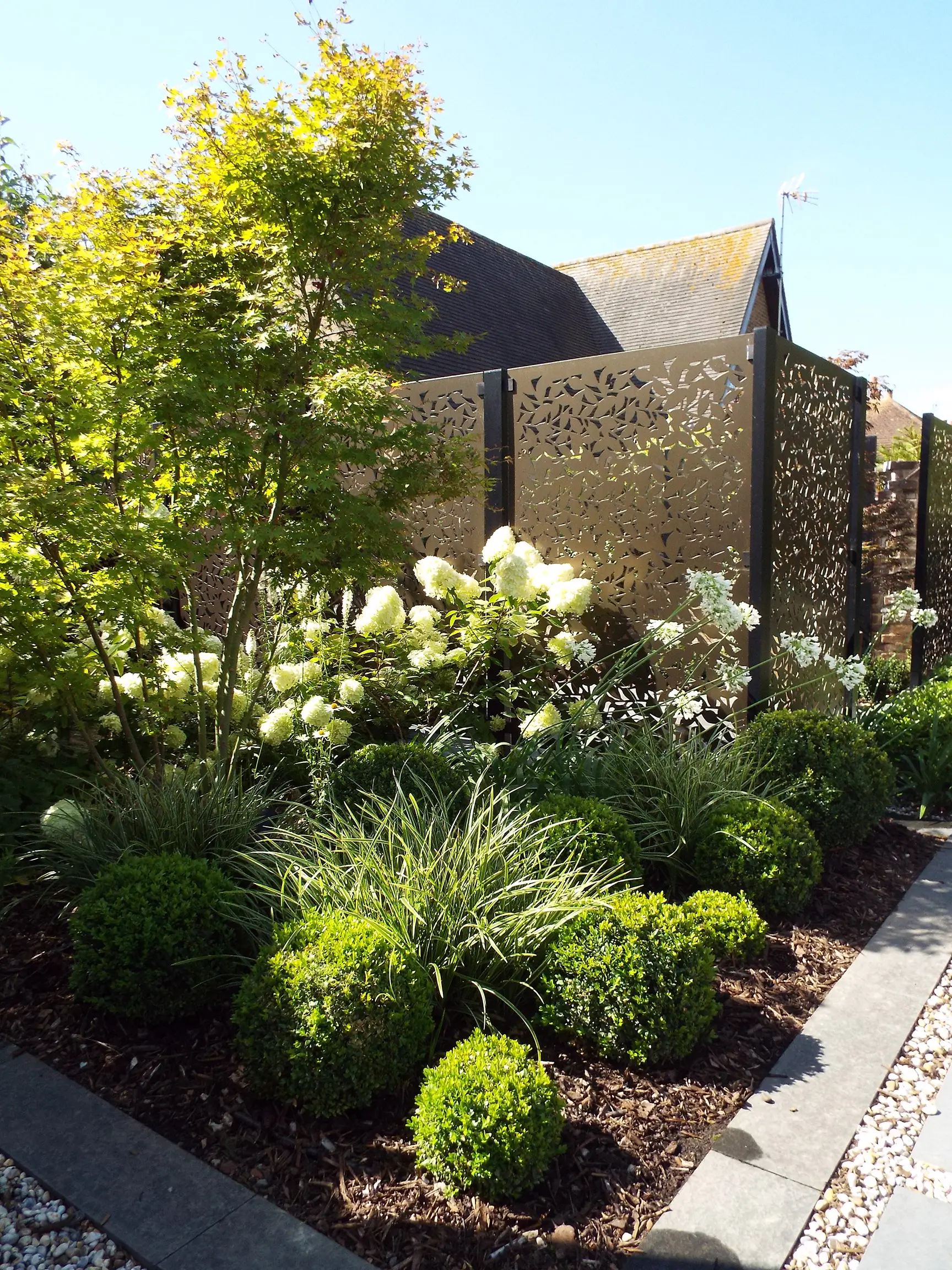 A vibrant garden filled with bushes and flowers, with a modern building in the background under a clear blue sky.