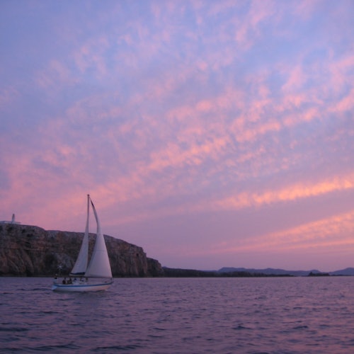 En segelbåt på lugnt vatten vid solnedgången, med en stenig kustlinje och en rosa och lila himmel med spridda moln.