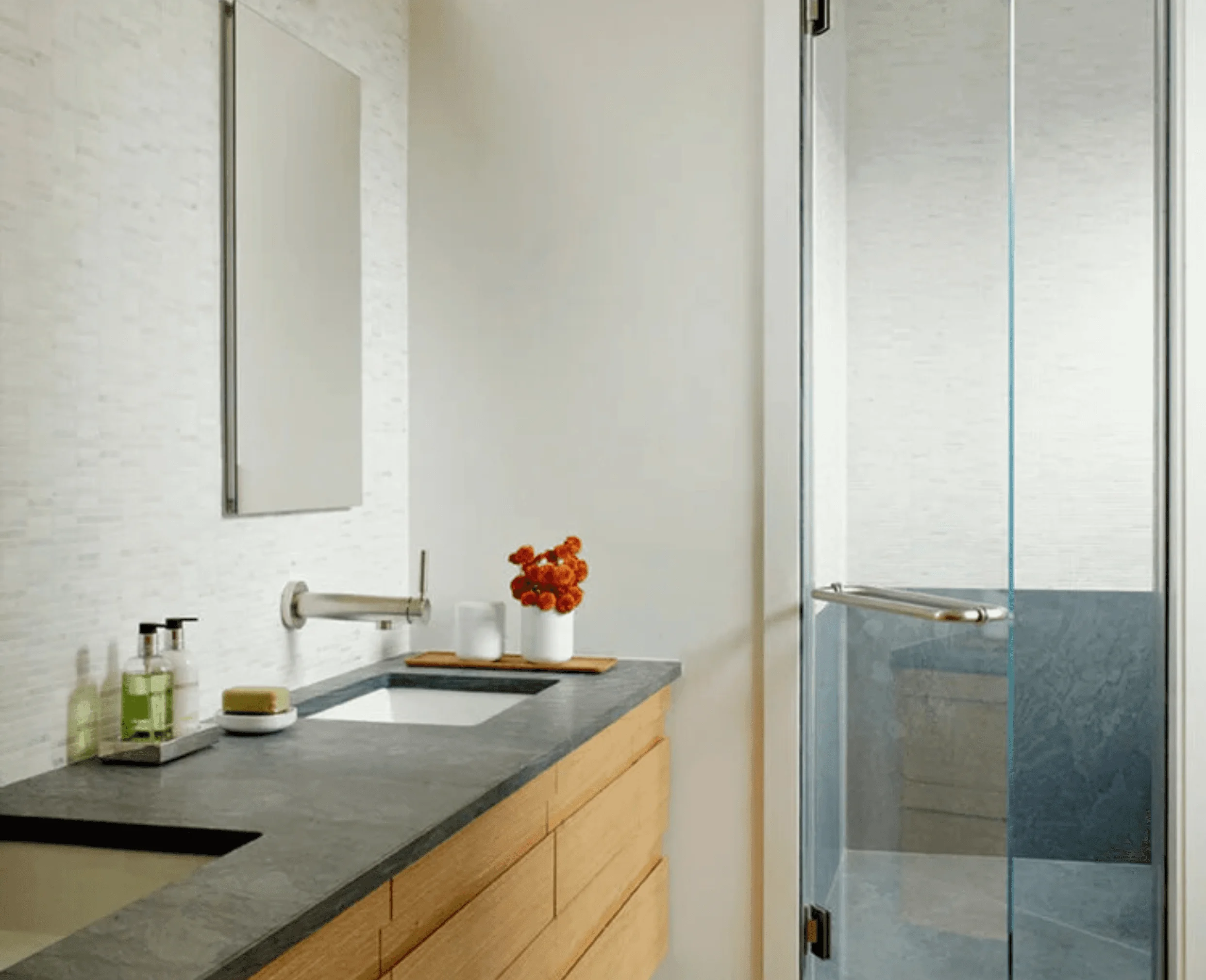 A bathroom vanity in honed dark stone and light wood cabinetry, beside a glass-enclosed shower