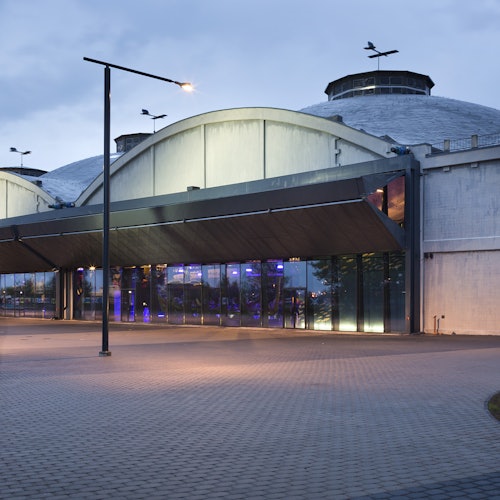 Modern épület íves tetővel, nagy üveg bejáratával és megvilágított sétányával, amely egy alkonyi égbolt előtt áll.