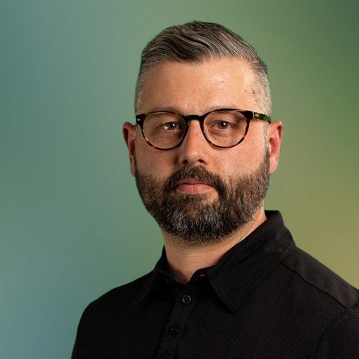 A headshot portrait of a white man with close cropped "salt and pepper" hair, glasses, a trimmed beard, and a black collared shirt, with a serious expression