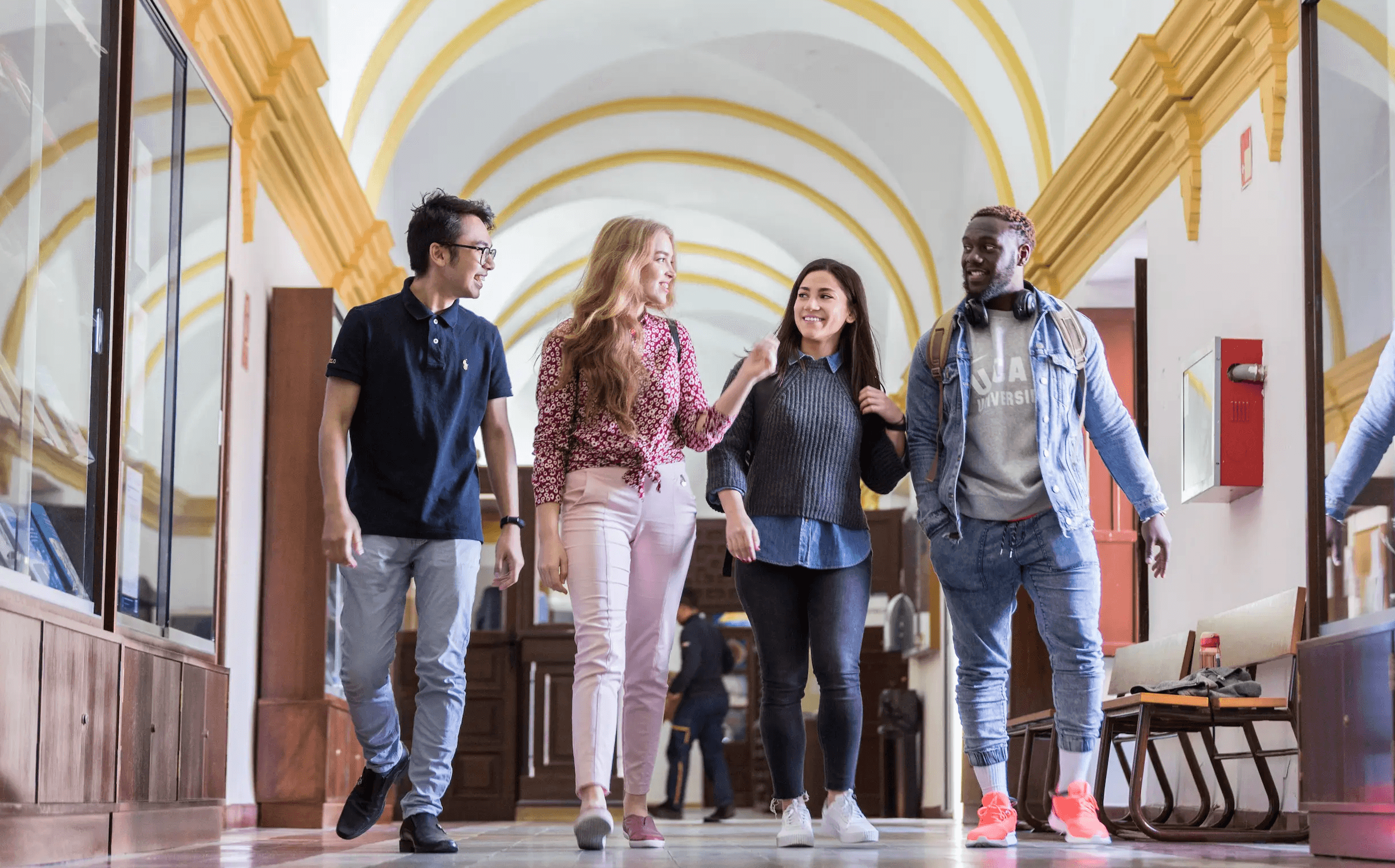 A diverse group of five students happily walking together outside a university campus, carrying books and backpacks among lush green trees and under a clear sky, reflecting a vibrant student life at UCAM Mumbai.