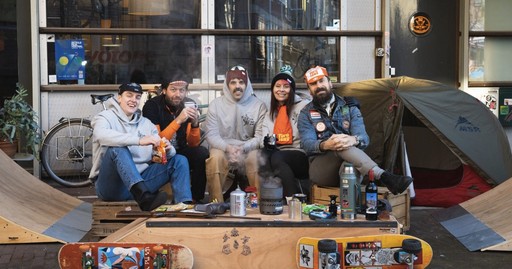 Six people sitting on a makeshift couch outdoors, enjoying drinks and snacks. Skateboards and a tent are nearby.