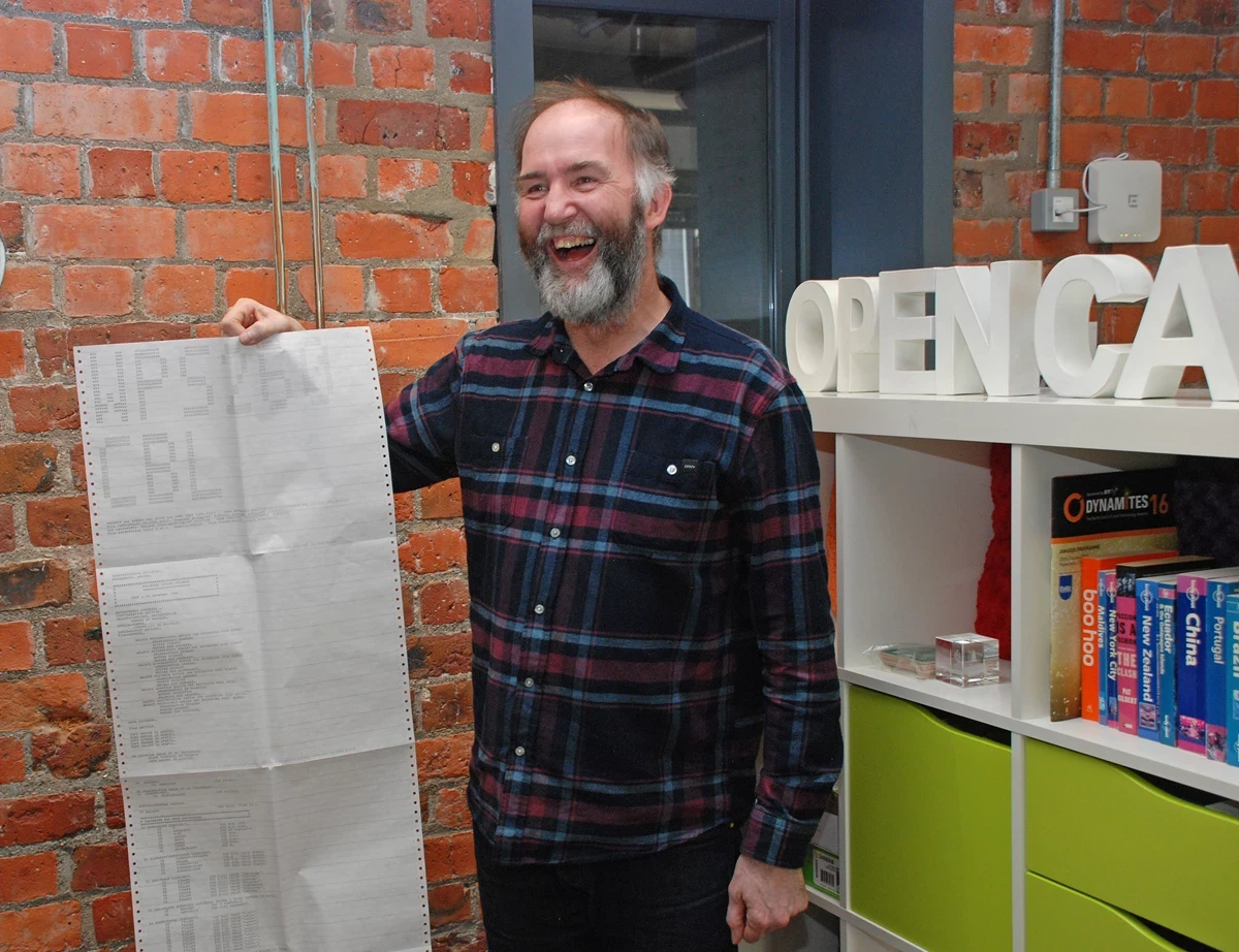 A person stands indoors beside a brick wall, holding up a long sheet of continuous computer printout paper. A shelf displaying books and large white letters spelling part of ‘OPENCAS…’ sits to the right.