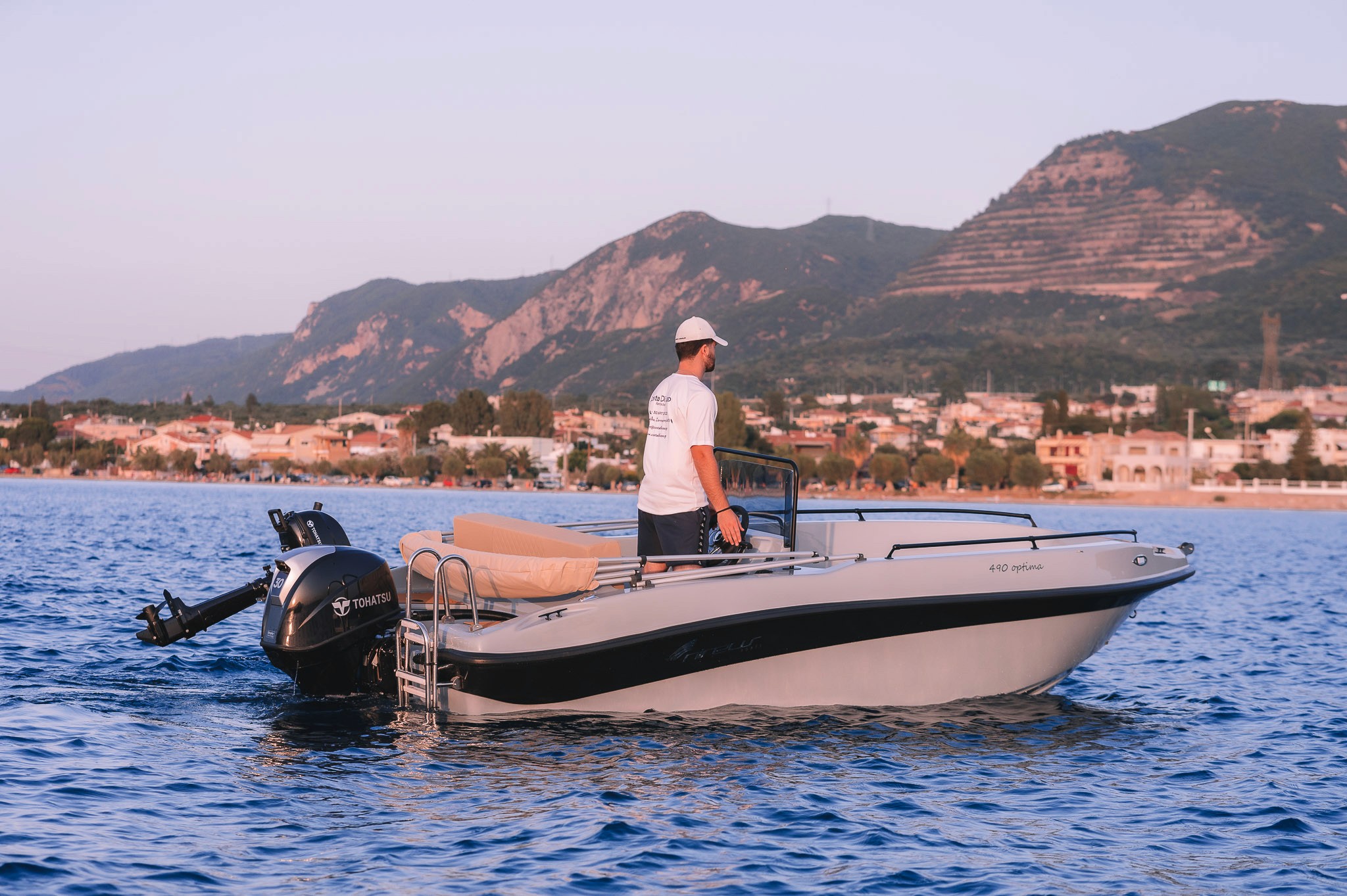 White speedboat with Tohatsu outboard motor anchored in calm blue waters, with captain standing at helm against backdrop of Greek coastal town and mountains at golden hour.