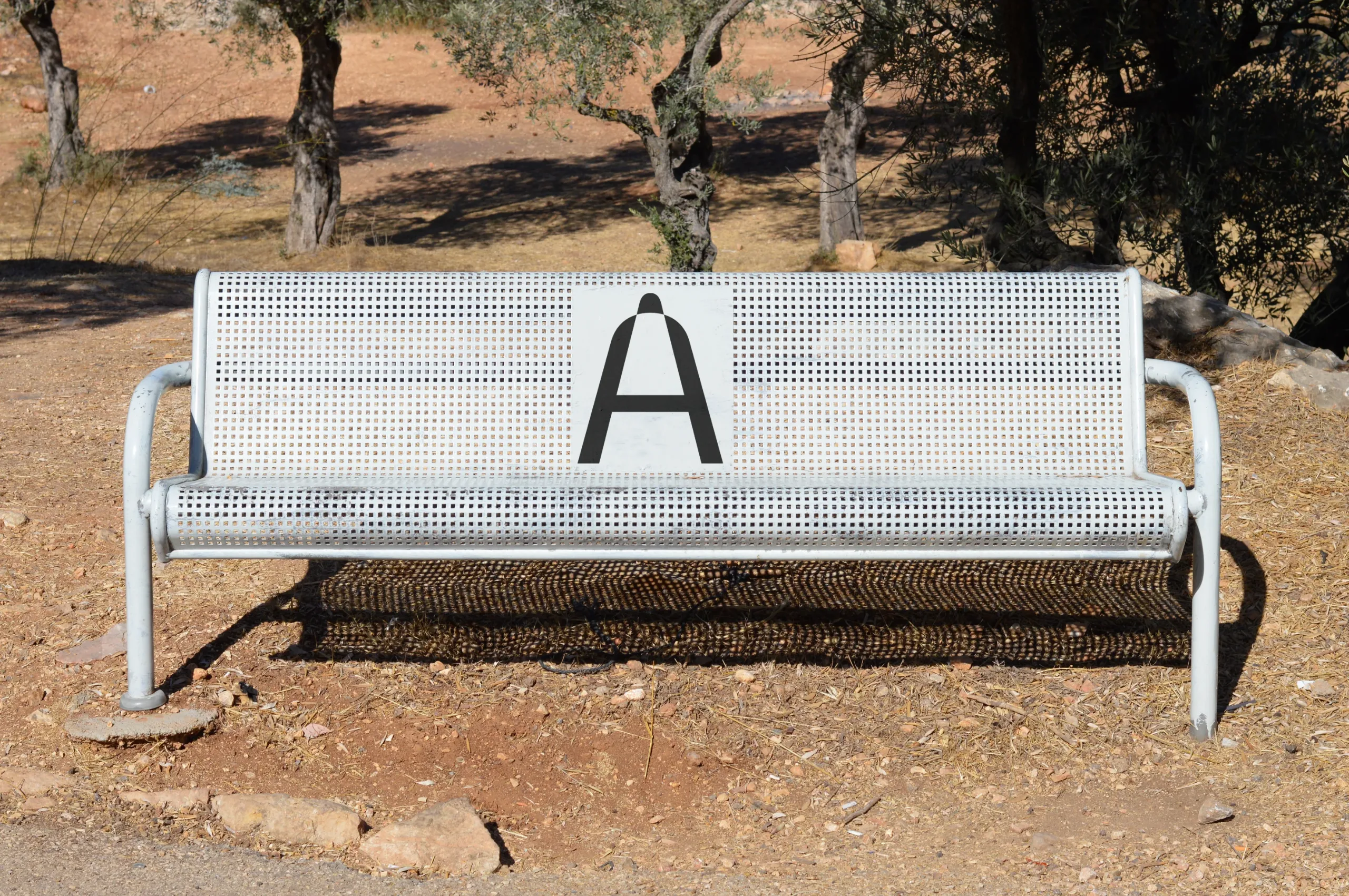 Outdoor bench mockup featuring a bright blue poster with four black circular designs. The bench has a perforated metal design, set in a sunlit rural environment with trees and dry earth in the background.