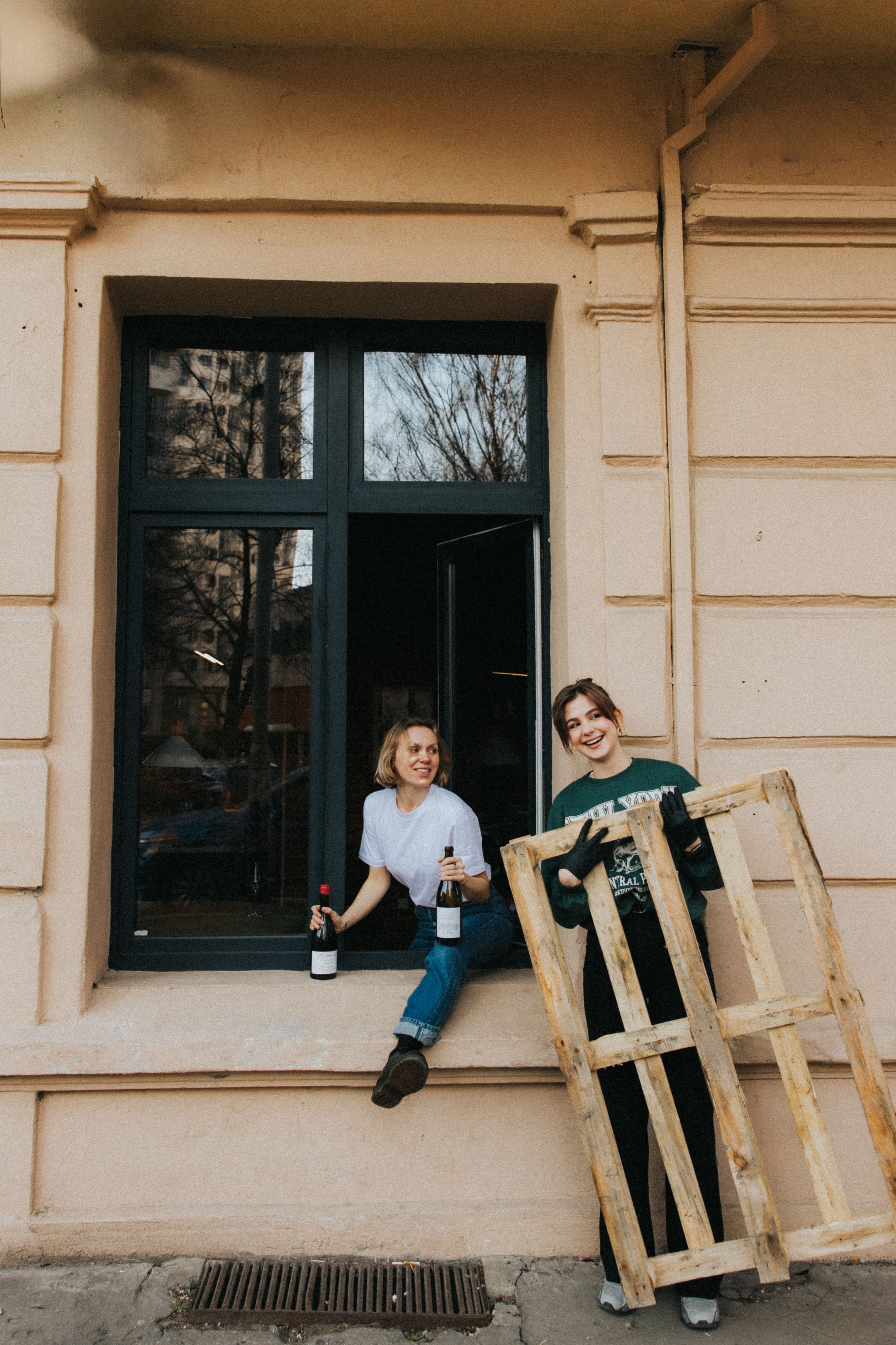 Dwie osoby przy oknie restauracji. Jedna siedzi na parapecie trzymając wino, podczas gdy druga stoi trzymając drewnianą paletę, oboje uśmiechają się ciepło.