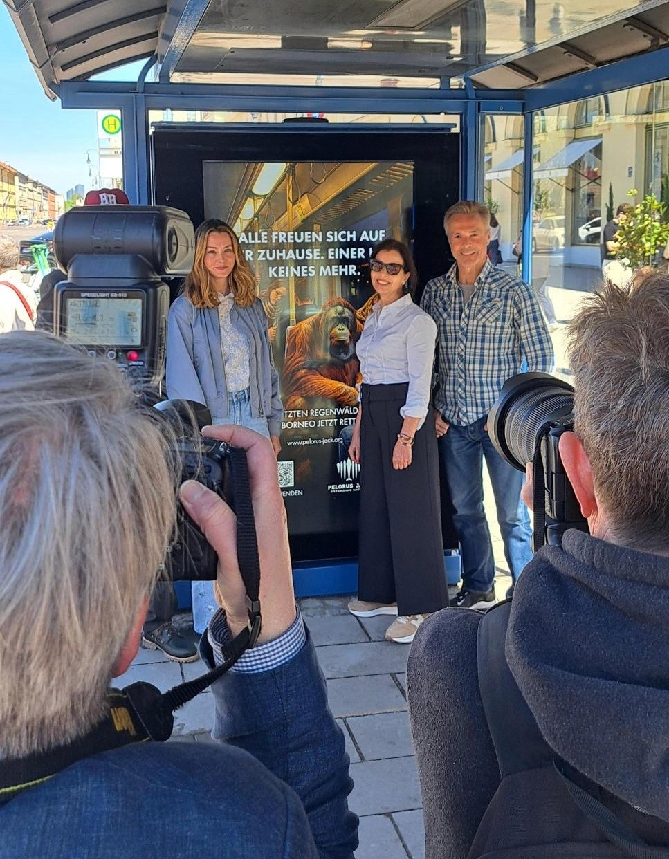 Larissa Theile, Nadja und Hannes Jaenicke posieren vor dem Kampagnenmotiv an einer Bushaltestelle in München, während Fotografen Bilder aufnehmen.