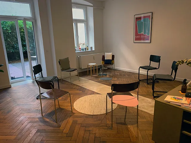 Ein gemütlicher Raum mit sieben Stühlen, die in einem Kreis auf einem hölzernen Fischgrätboden angeordnet sind. Sanftes Licht und ein Teppich schaffen eine einladende, entspannte Atmosphäre.