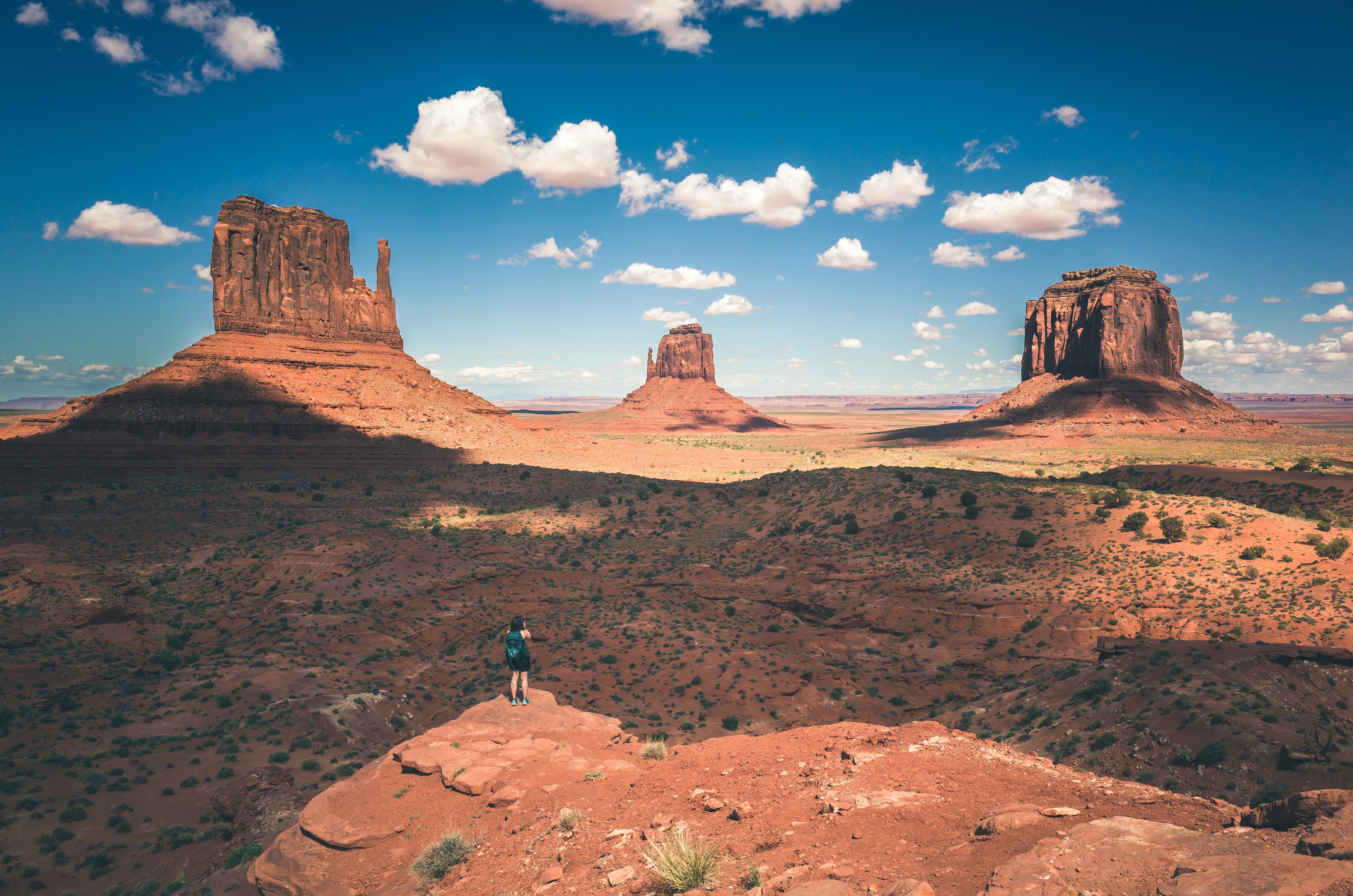 The West and East Mitten Buttes and Merrick Butte rising from the red desert floor at Monument Valley, with a lone visitor standing on a rock ledge in the foreground for scale.