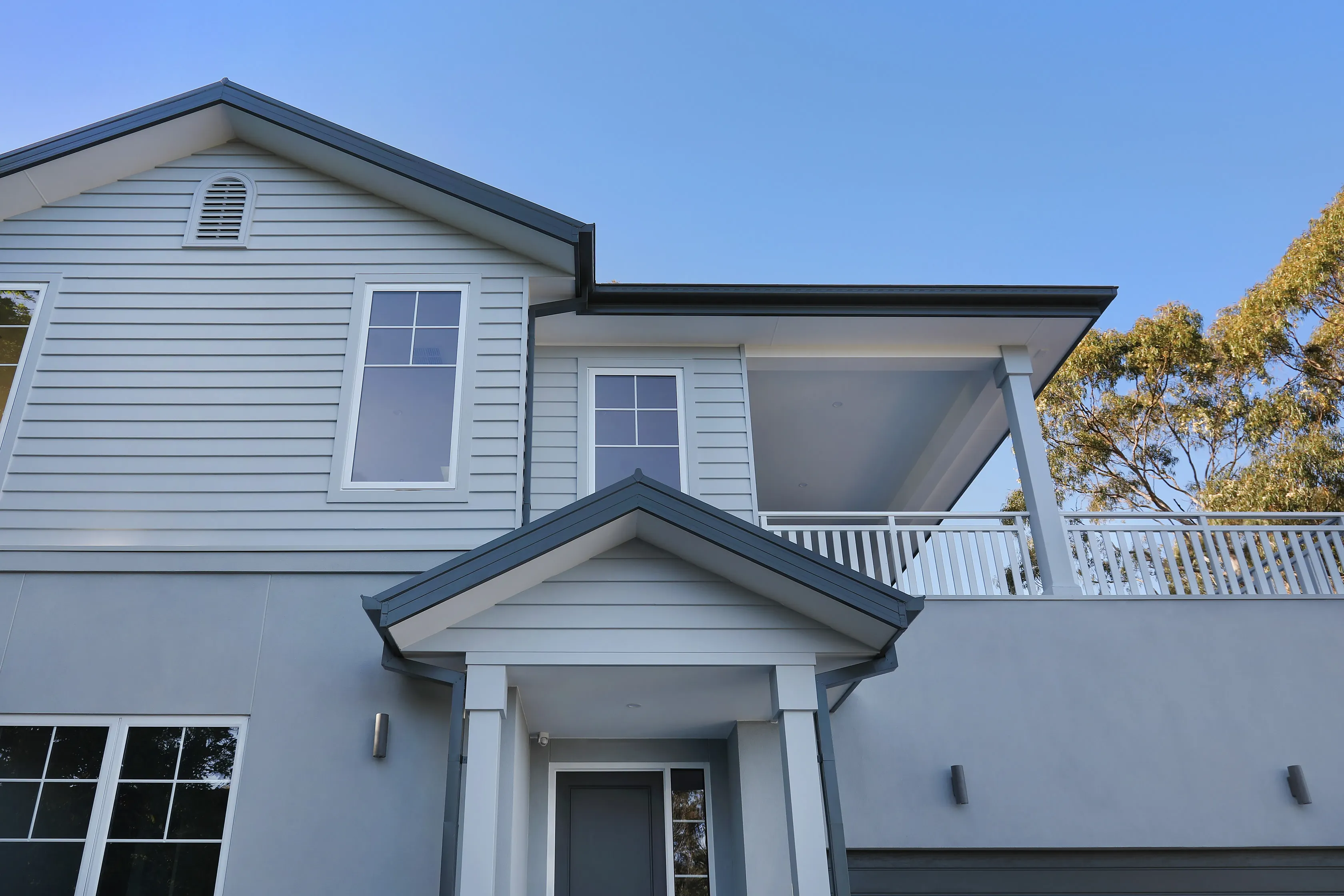 Upper facade of the Eltham Residence in pale weatherboard with white-trimmed windows, a gabled entry portico, columned balcony with white balustrade and gum trees against blue sky