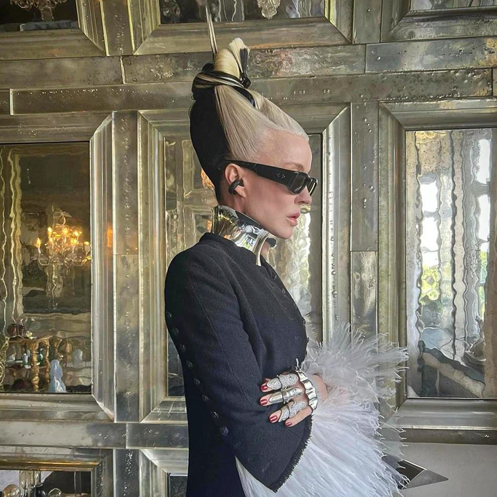 Profile portrait of style icon Daphne Guinness styled with a high-sculpted updo, dark sunglasses, and a feather-cuffed jacket, wearing a polished sterling silver neck collar and diamond-encrusted finger armor by designer Shaun Leane.