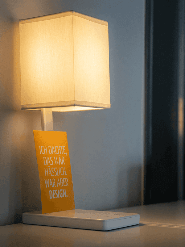 Tischlampe mit rechteckigem Lampenschirm und gelber Karte mit der Aufschrift 'ICH DACHTE, DAS WÄR HÄSSLICH. WAR ABER DESIGN.'