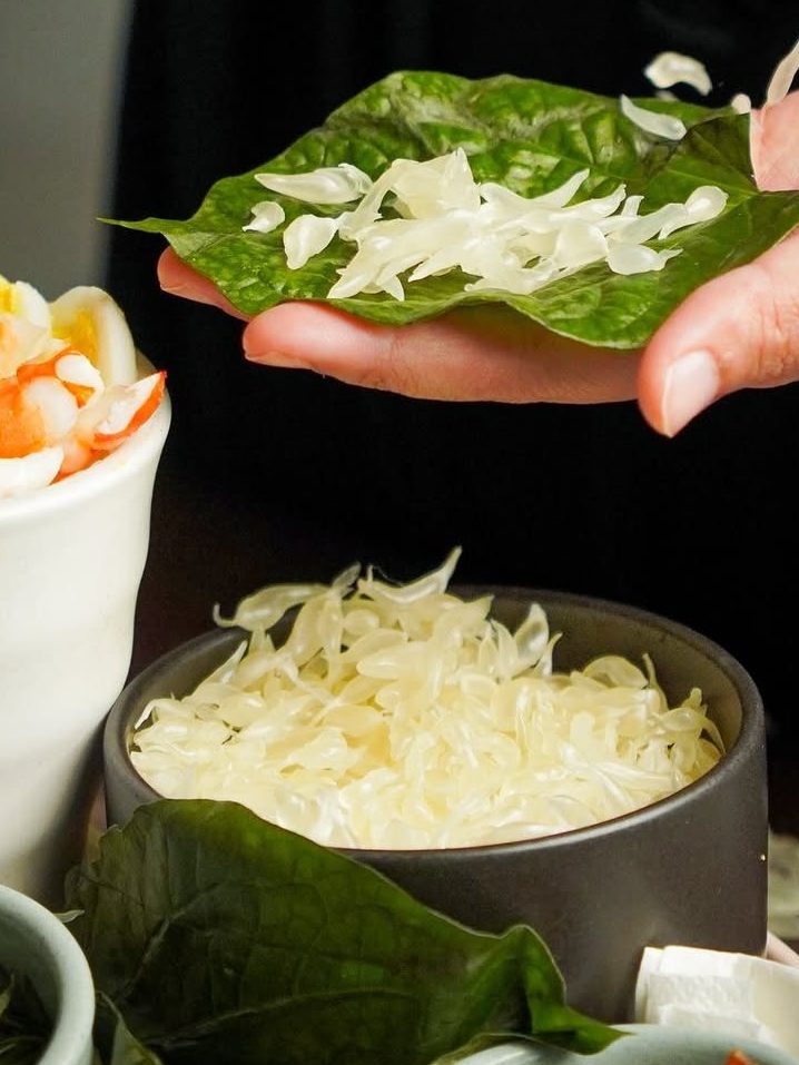 A hand holding a green leaf with shredded vegetables, next to bowls of shrimp, additional shredded vegetables, and various small bowls with condiments.