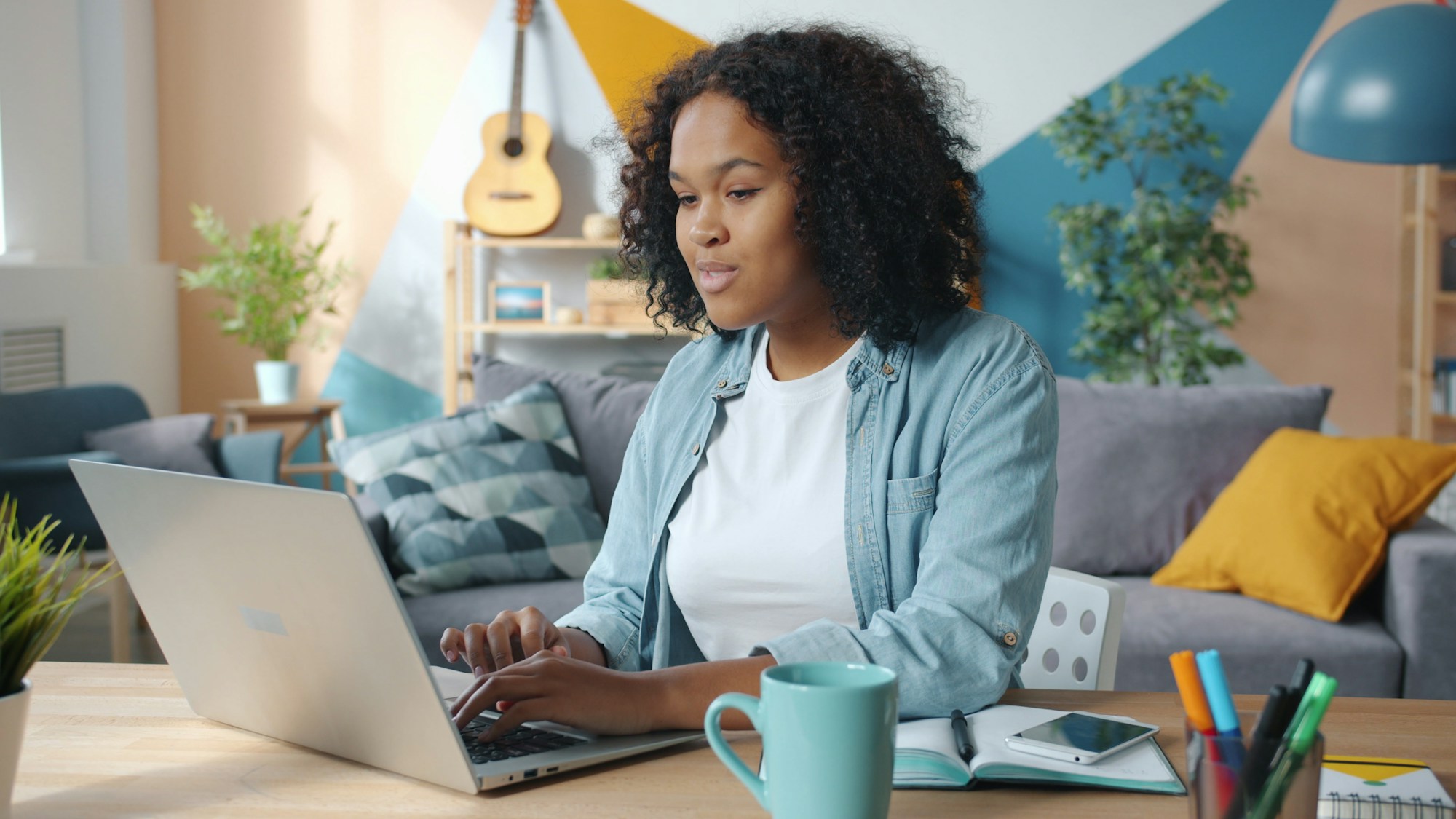 A woman working on a laptop at a desk in a bright living room, with a notebook and a mug beside her